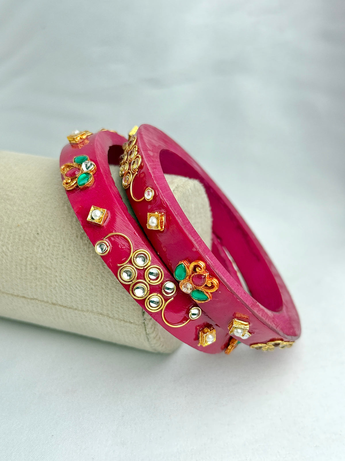 Two bright pink bangles with gold-toned floral and rhinestone embellishments, displayed on a cream-colored jewelry stand against a light gray background.