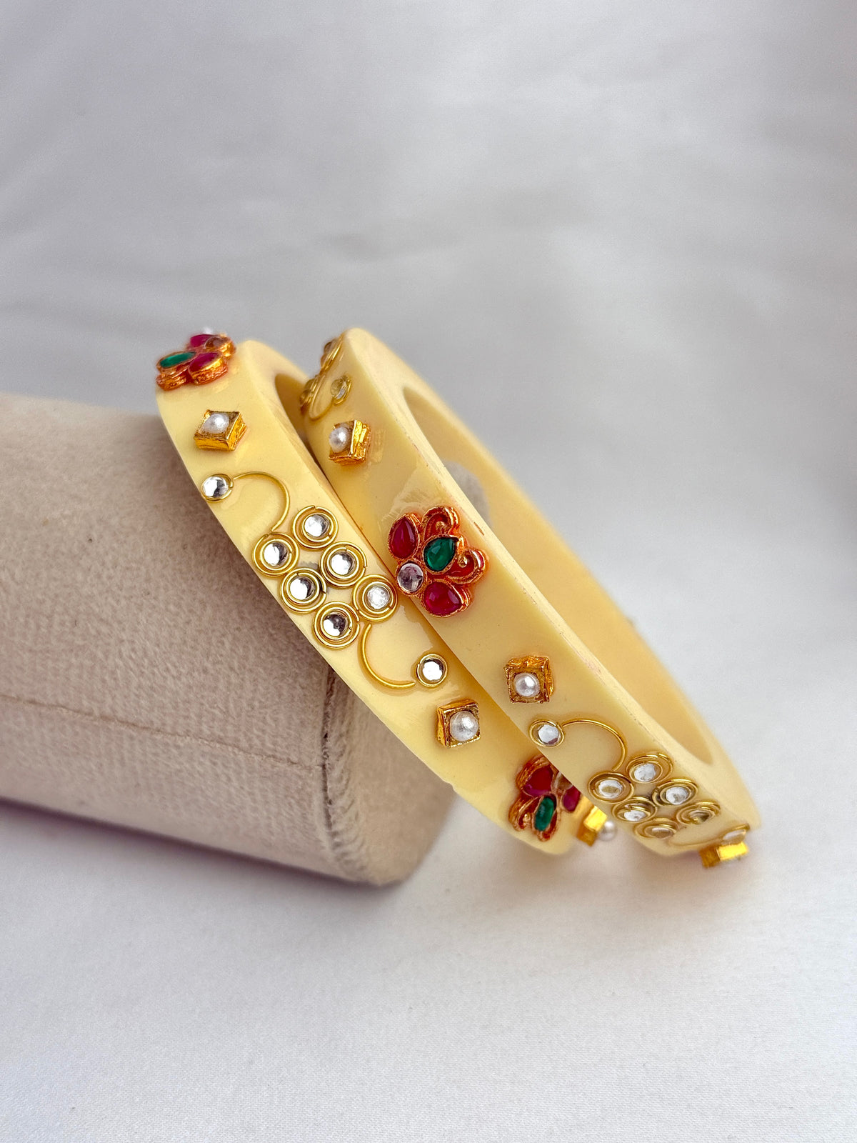Two yellow bangles with gold-toned floral and circular embellishments, decorated with small white stones and red and green enamel details, displayed on a beige cylindrical stand against a light gray background.