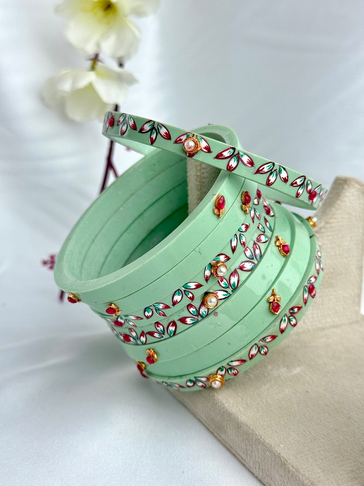 A stack of mint green bangles decorated with red, gold, and white floral patterns, displayed on a neutral surface with a soft-focus background of white flowers.