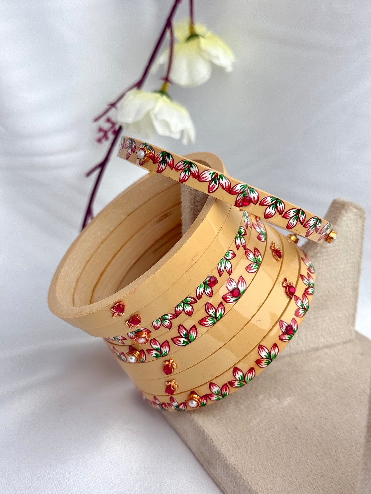 A stack of light beige wooden bangles decorated with red, green, and gold floral patterns, displayed on a neutral stand against a white fabric backdrop with white flowers in the background.