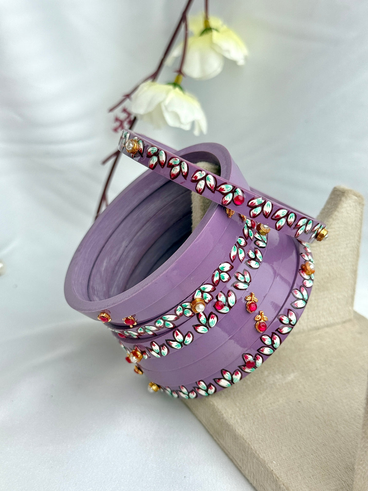 A lavender purple traditional Indian bindi bracelet adorned with colorful rhinestones, displayed on a beige jewelry stand with white flowers in the background.