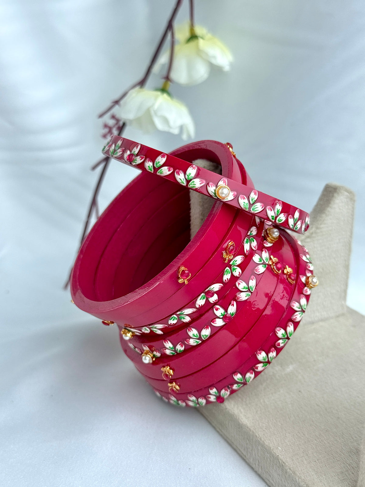 A stack of bright pink bangles decorated with white, green, and gold floral enameled details, displayed on a neutral stand against a soft white background with decorative white flowers.