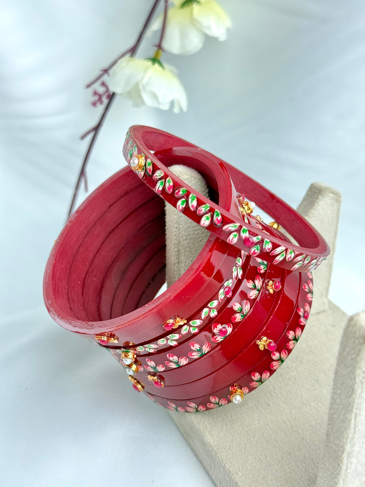 A stack of glossy red bangles decorated with small floral motifs in pink, green, and gold, displayed on a beige jewelry stand against a soft white background with a blurred flower stem.