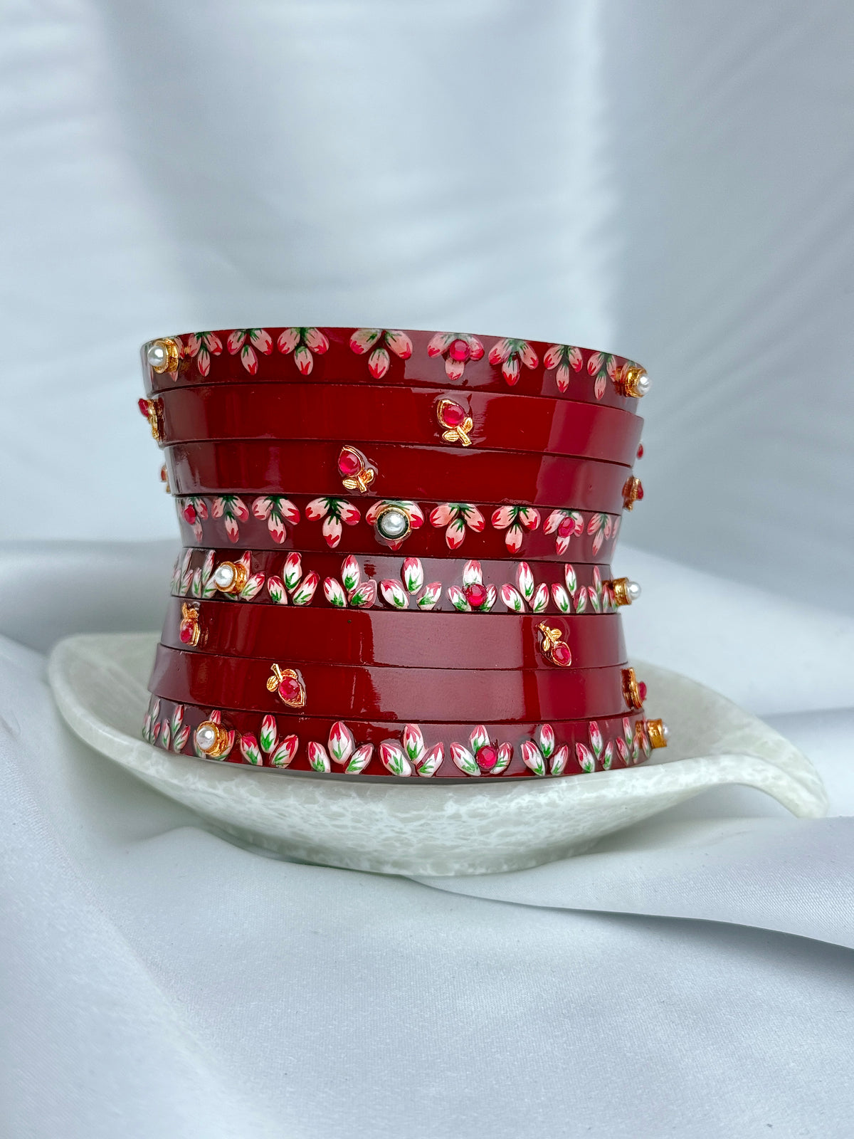 A stack of red bangles decorated with floral patterns and small gold-toned embellishments, displayed on a white ceramic dish against a soft white fabric background.