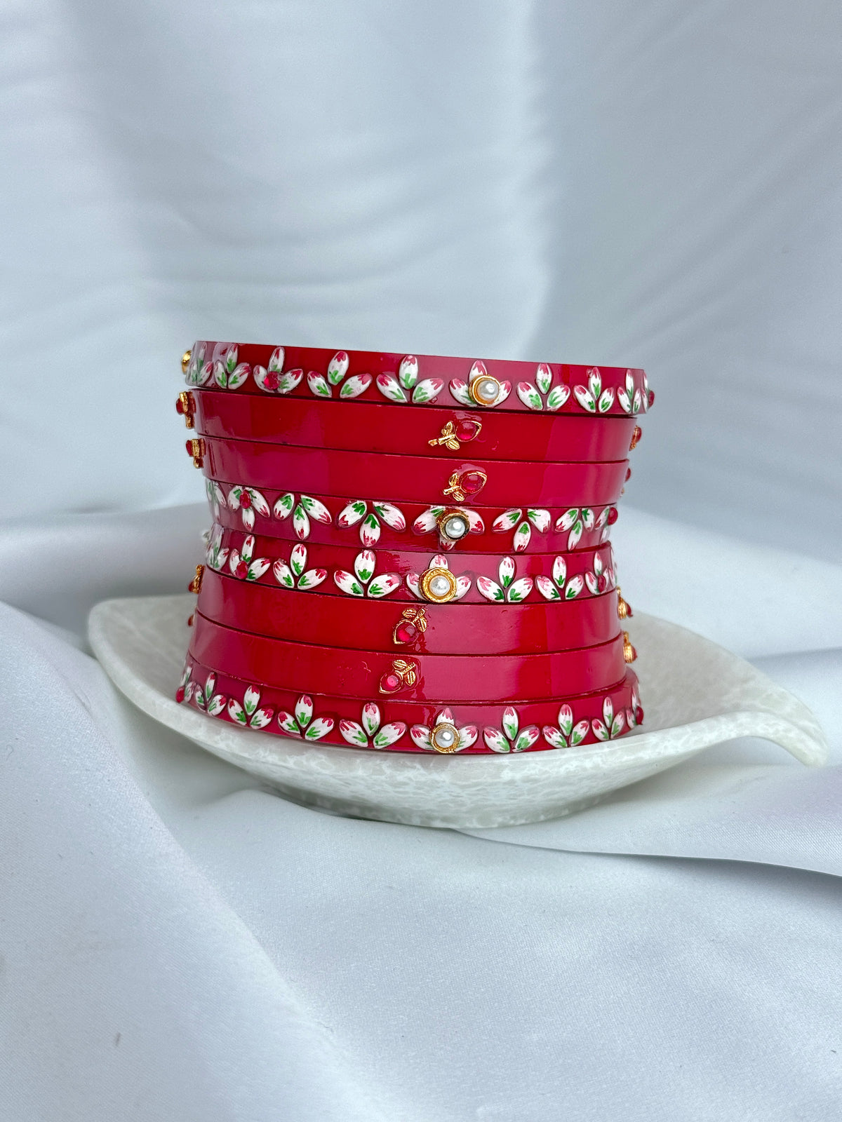 A stack of red bangles decorated with white and green floral patterns and gold accents, displayed on a white ceramic plate against a draped white fabric background.