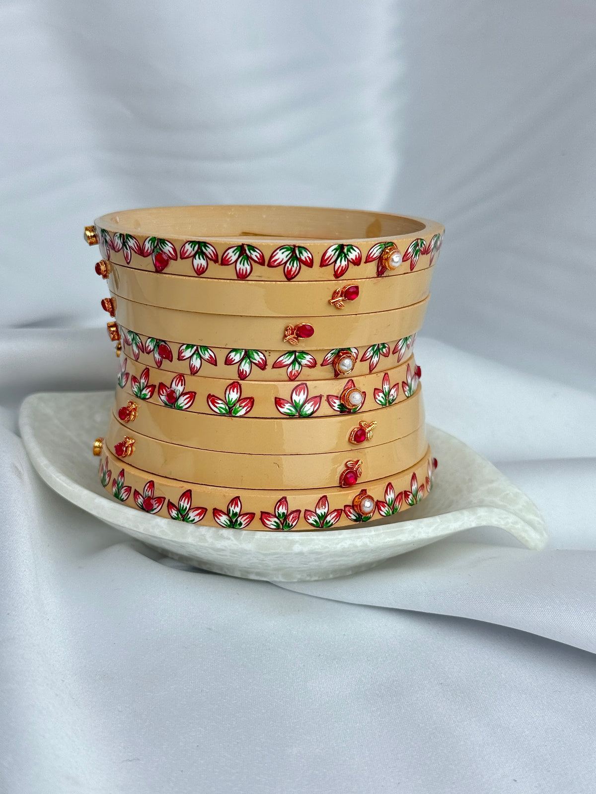 A stack of eight light beige bamboo bangles with red, green, and white floral patterns and small gold bead accents, displayed on a white ceramic dish against a draped white fabric background.