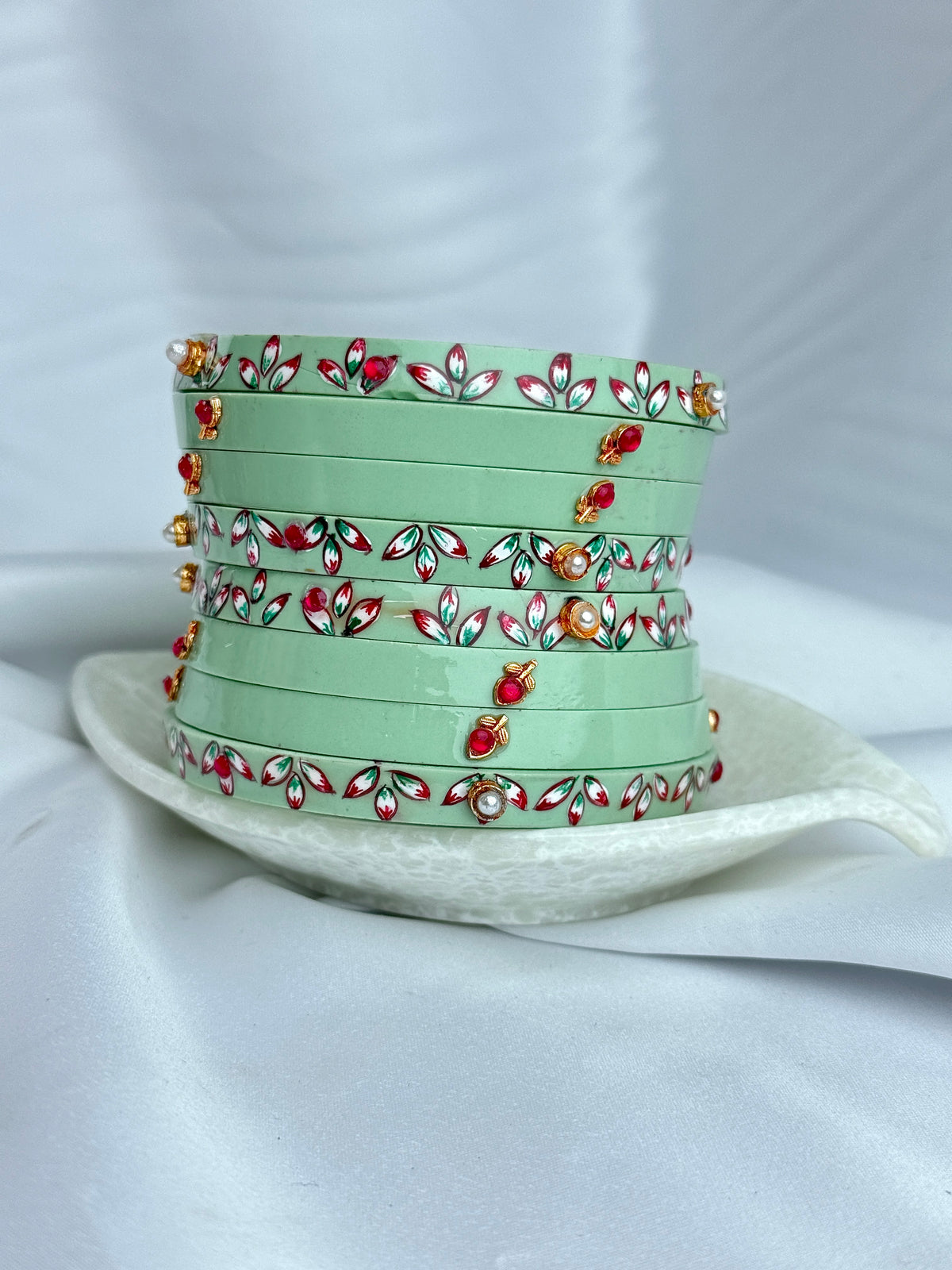 A stack of mint green bangles decorated with red, white, and green floral patterns and small gold-toned embellishments, displayed on a white ceramic plate against a white fabric backdrop.