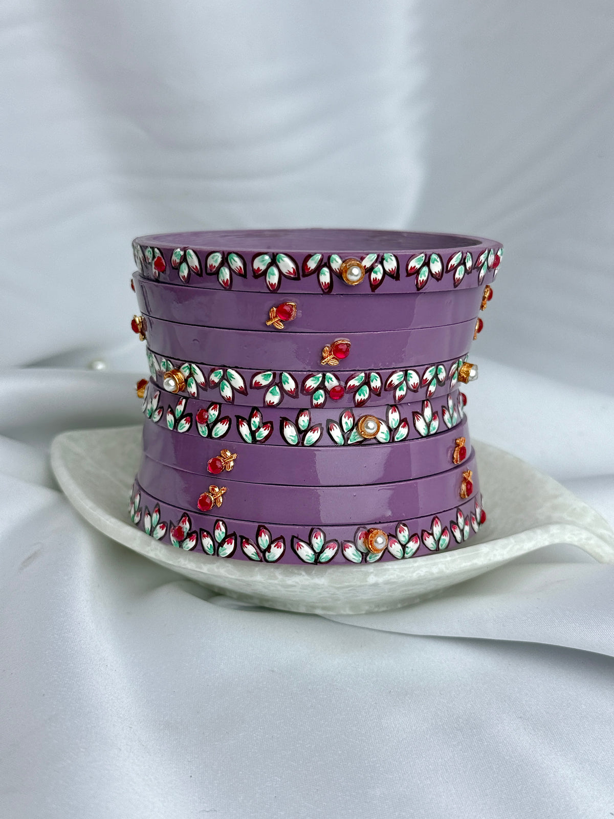 A stack of five purple cylindrical containers or boxes with decorative floral borders and small red and orange floral accents, displayed on a white dish against a white draped fabric background.
