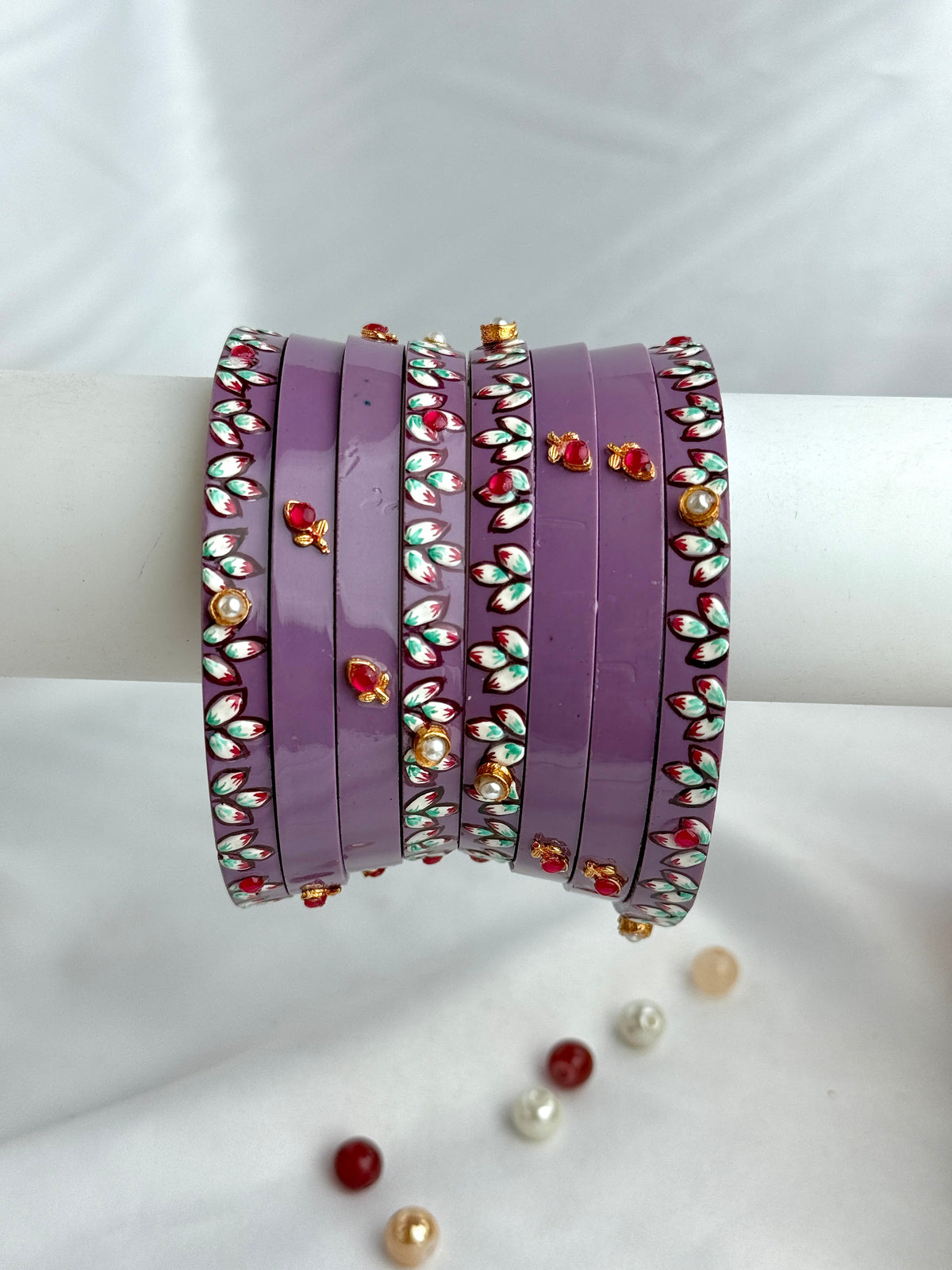 A stack of purple bangles adorned with floral enameled details in red, green, white, and gold, displayed on a white cylindrical stand against a white fabric background.