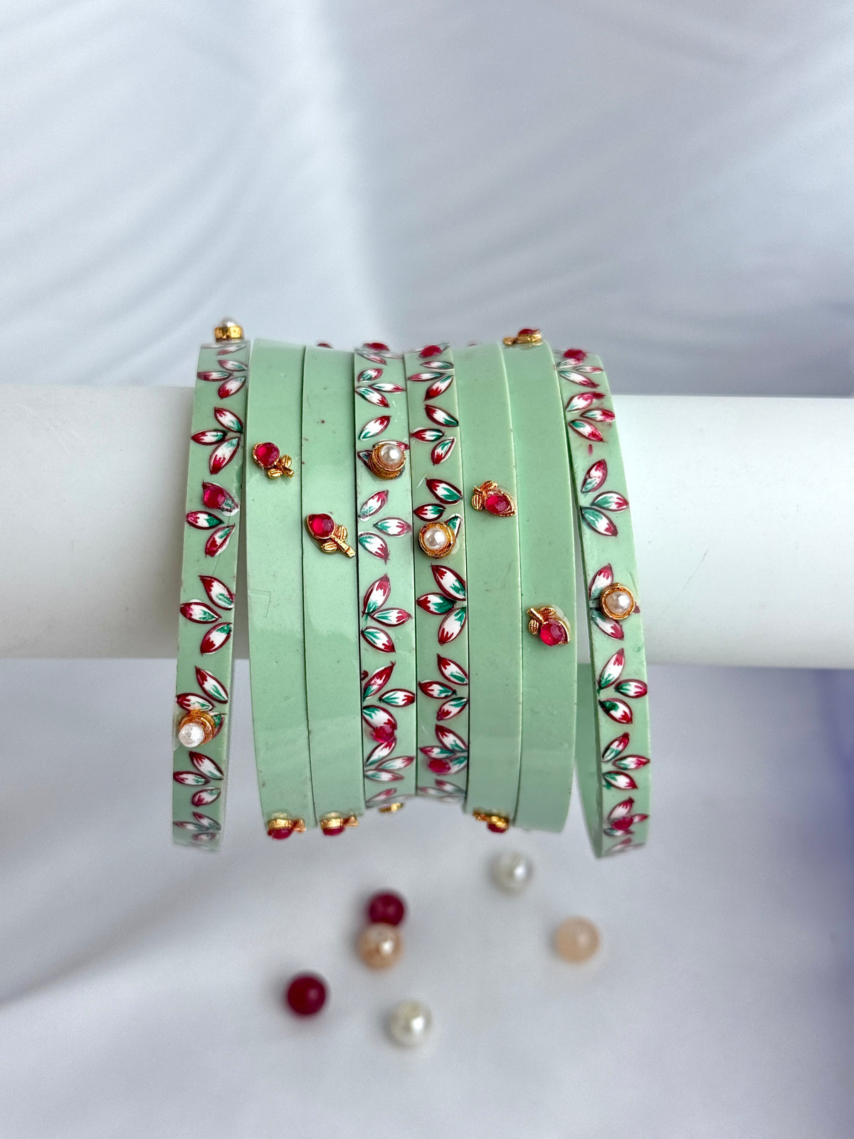 A set of mint green bangles decorated with red, white, and gold floral patterns and small pearl-like beads, displayed on a white cylindrical stand against a white fabric background.