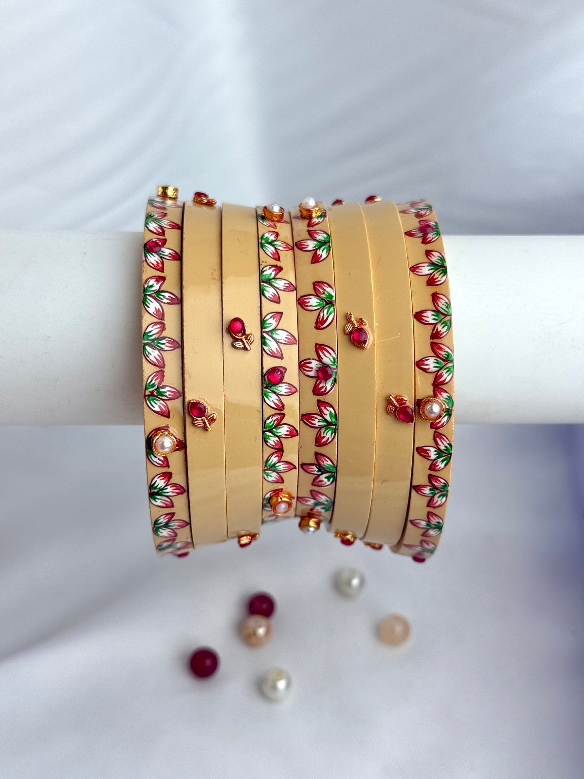 A stack of gold-toned bangles decorated with red and green floral patterns and small pearl-like accents, displayed on a white cylindrical stand against a white fabric background.