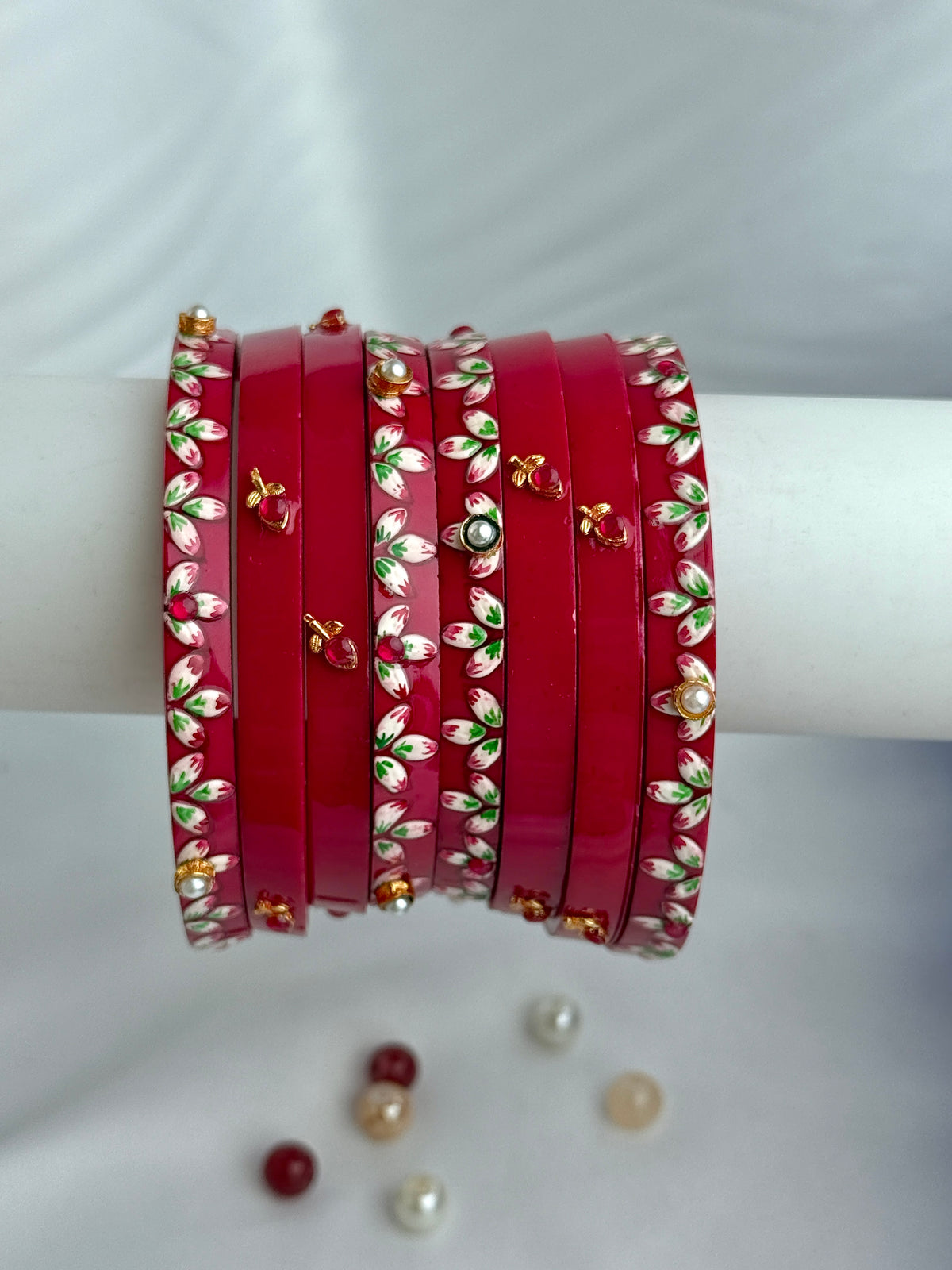 A stack of red bangles with white floral patterns, green leaf accents, and small gold and red decorative details, displayed on a white cylindrical stand against a white background.