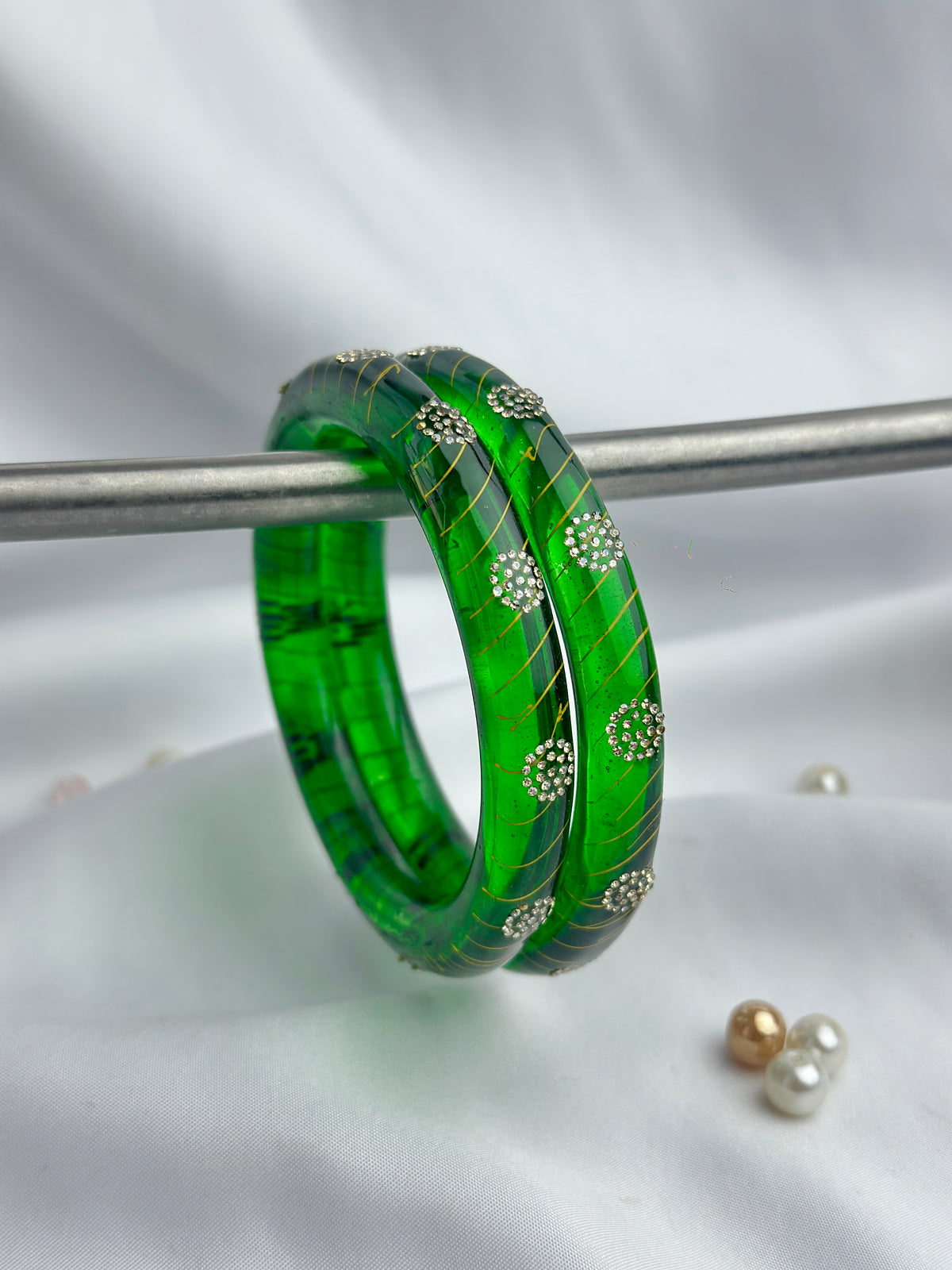 A pair of green translucent bangles with gold diagonal stripes and silver floral motifs, displayed on a metal stand against a white fabric background.