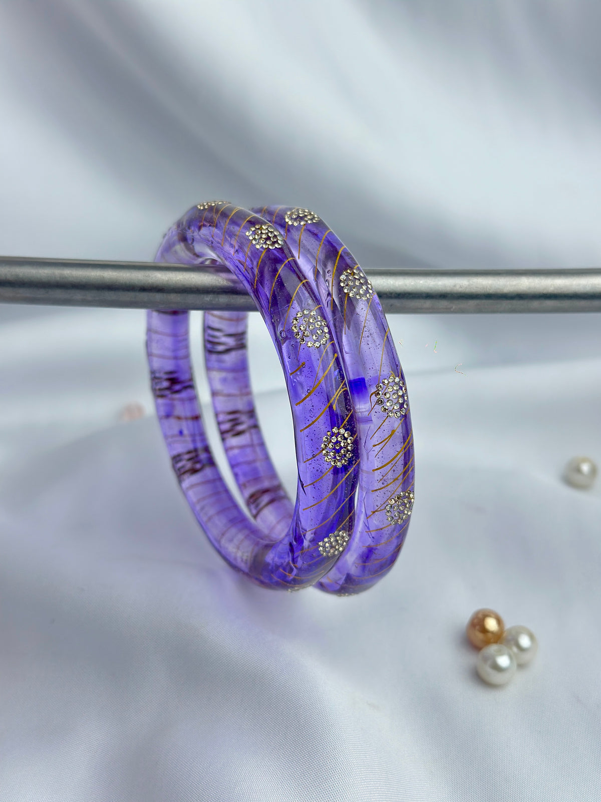 A purple translucent bangle bracelet with gold swirls and silver floral accents, displayed on a metal stand against a white fabric background.
