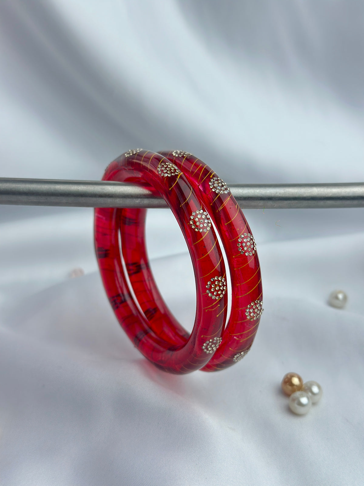 A pair of red bangles with gold-toned circular embellishments, displayed on a metal stand against a white fabric backdrop.