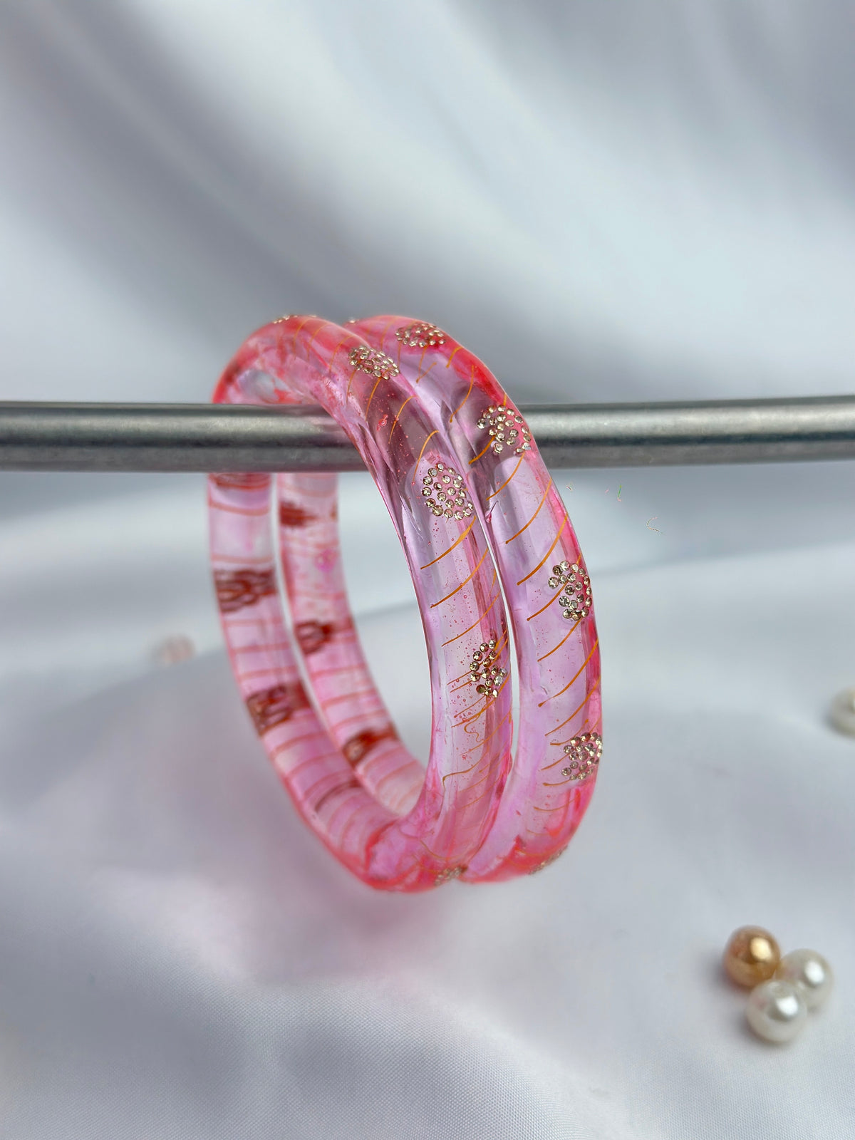 A pair of translucent pink bangles with gold floral details and red striped accents, displayed on a metal stand against a white fabric background.