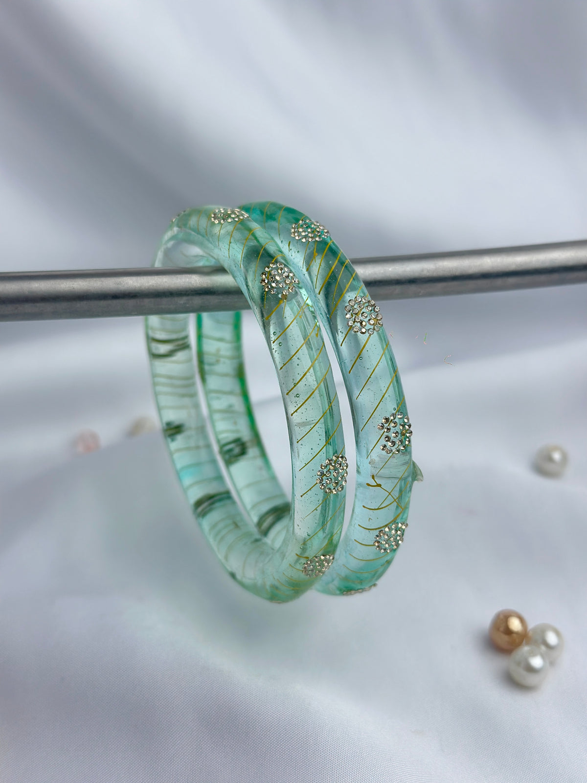 Two light green glass bangles with gold leaf swirls and small gold floral accents, displayed on a silver stand against a white fabric background.
