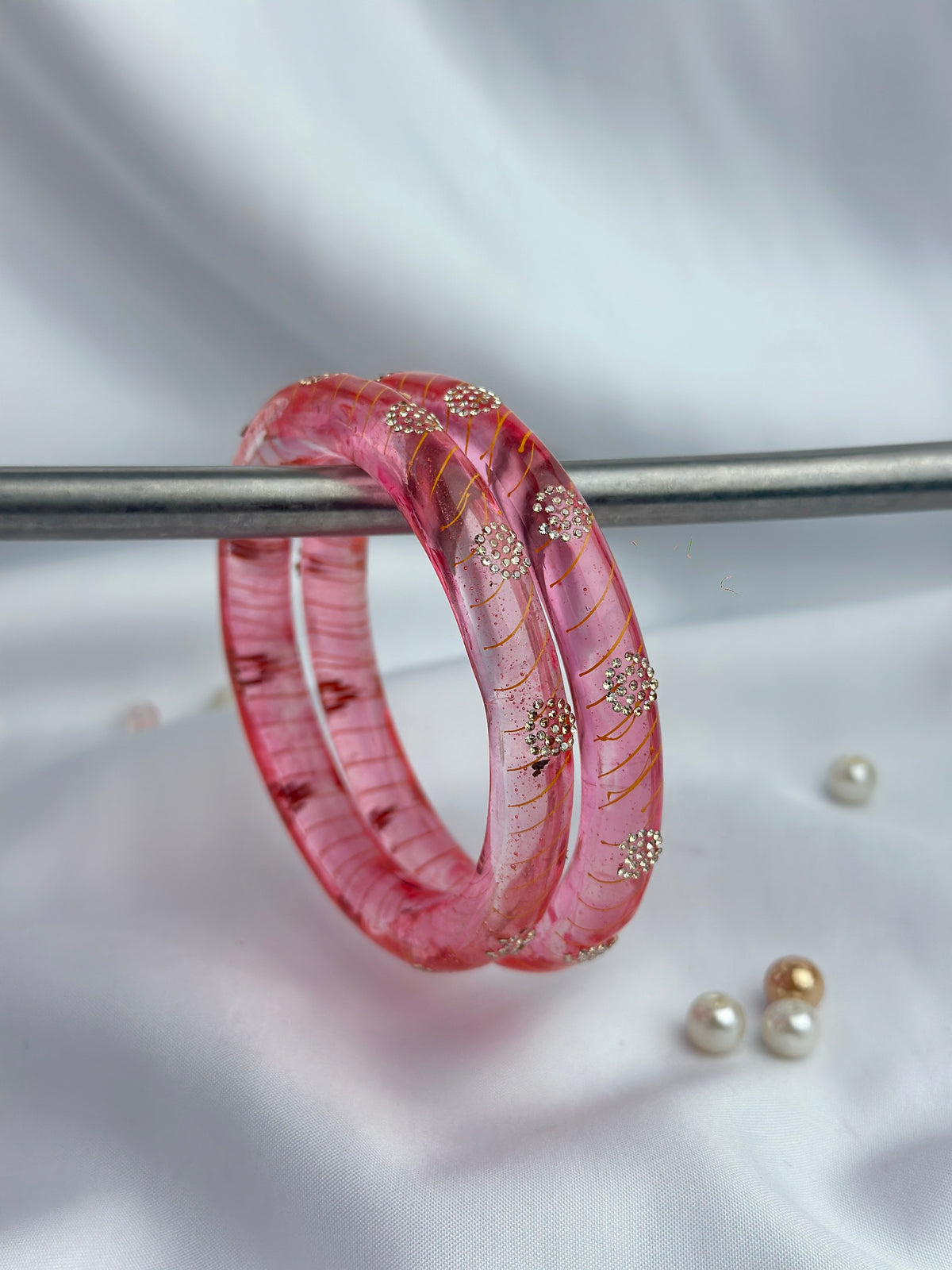 A pair of pink translucent bangles with gold floral details, resting on a silver rod against a white satin background with small pearls nearby.