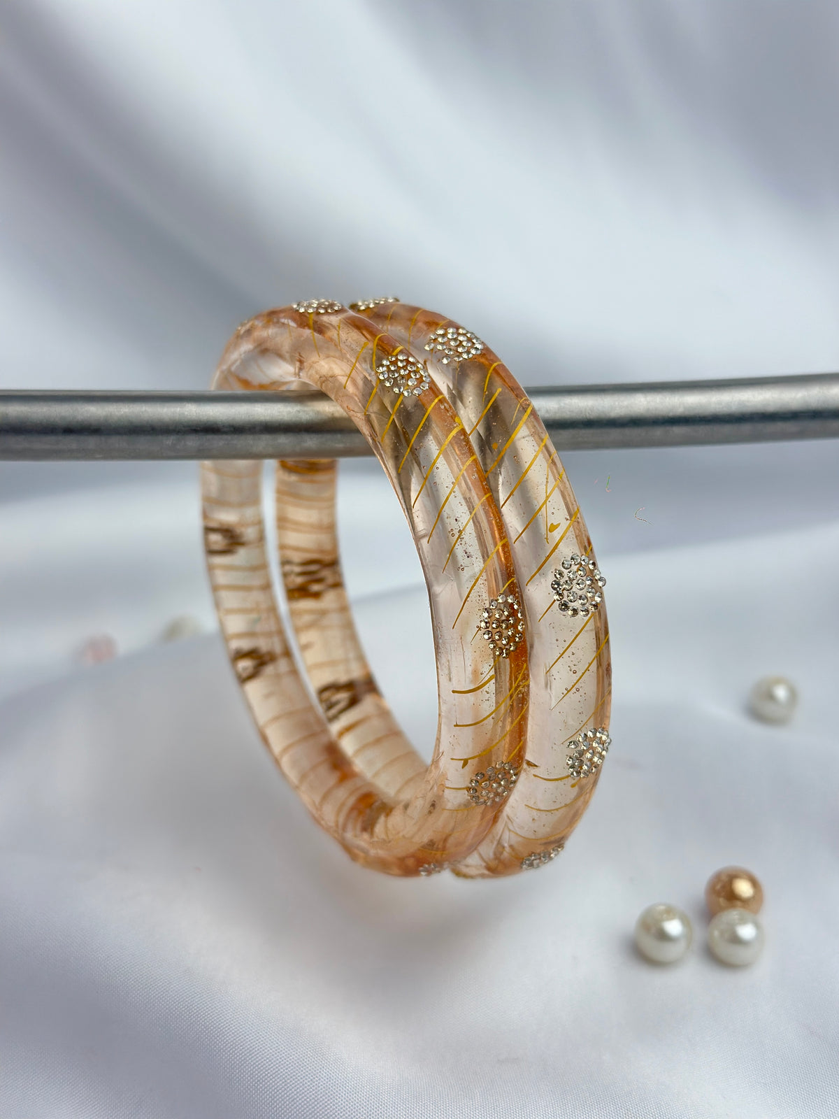 A transparent peach-colored resin bangle bracelet with gold glitter lines and silver rhinestone accents, displayed on a metal stand against a white satin background.