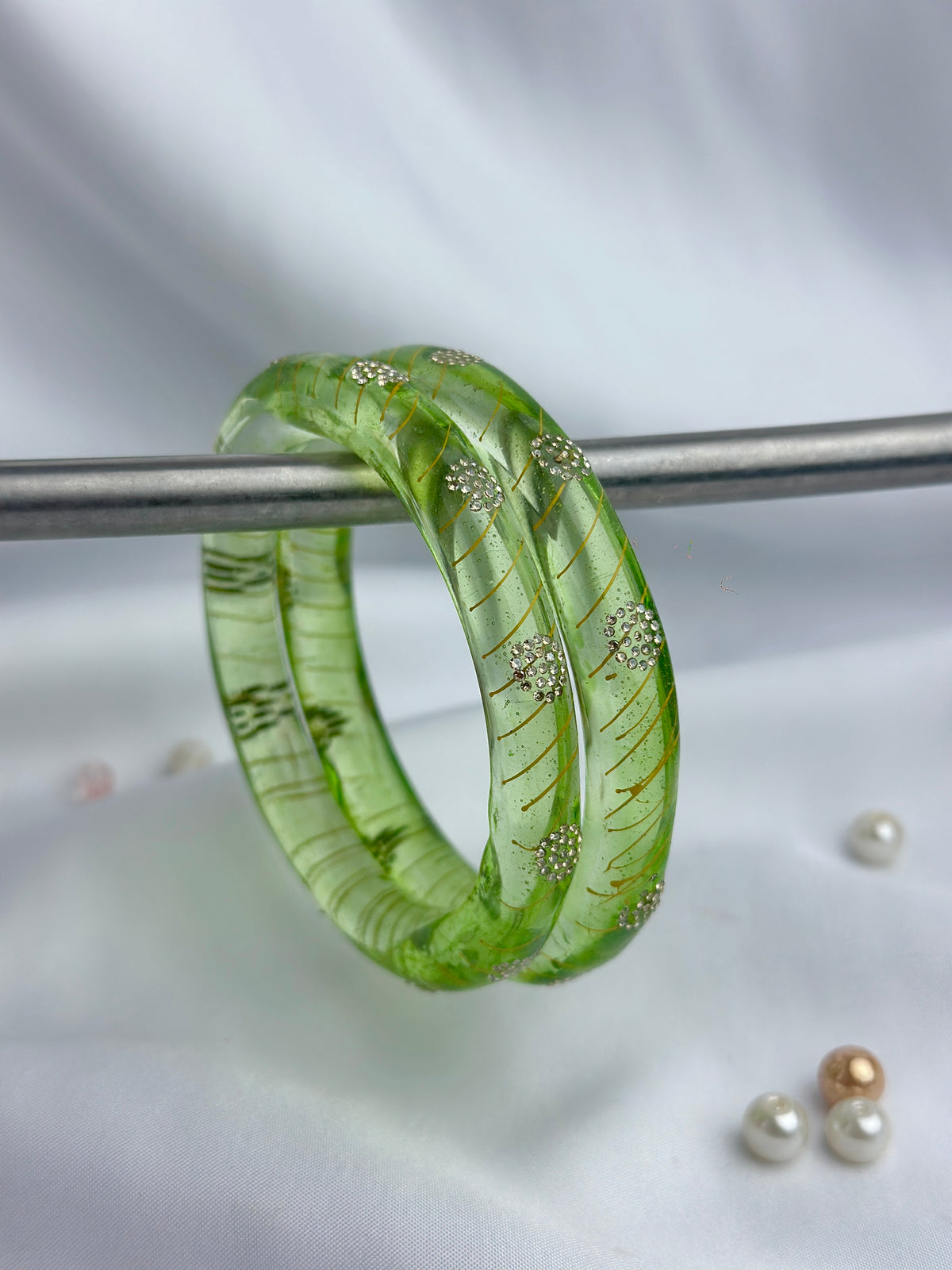 A translucent green glass bangle bracelet with gold leaf and small crystal-like inclusions, resting on a metal rod against a soft white fabric background.