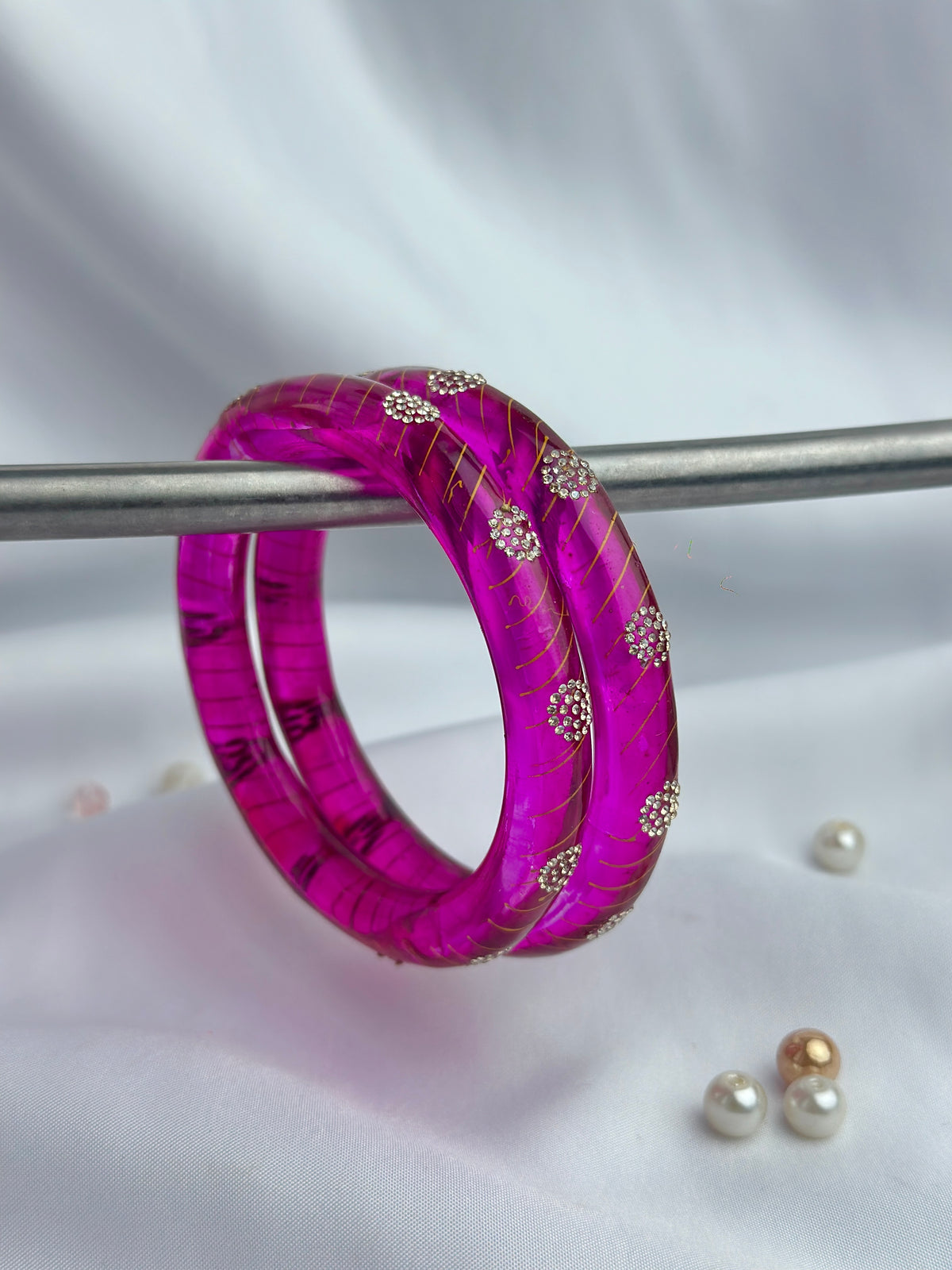 A magenta pink resin bangle bracelet with gold-toned metallic veins and silver floral crystal accents, displayed on a metal stand against a white draped fabric background.