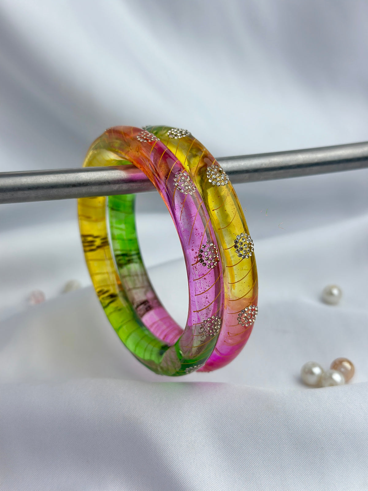 A translucent bangle bracelet with a vibrant rainbow gradient of pink, yellow, green, and orange, decorated with small gold floral motifs and rhinestone accents, displayed on a silver stand against a white satin background.