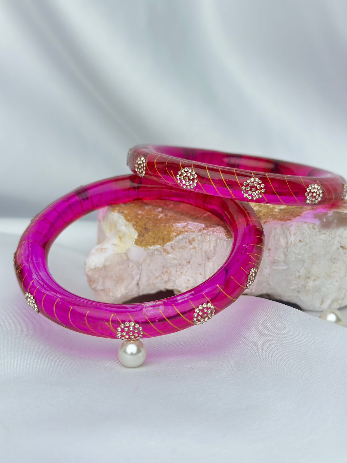Two translucent hot pink bangles with gold veining and small silver floral accents, displayed on a white fabric backdrop with a decorative stone prop.