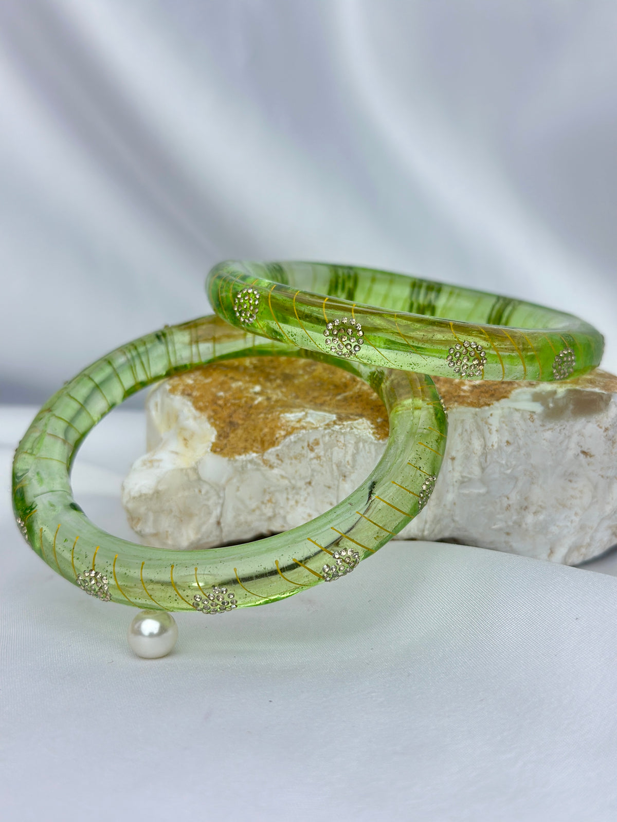 Two translucent green bangles with gold striped detailing and small gold floral accents, displayed on a white surface with a textured stone prop and a pearl.