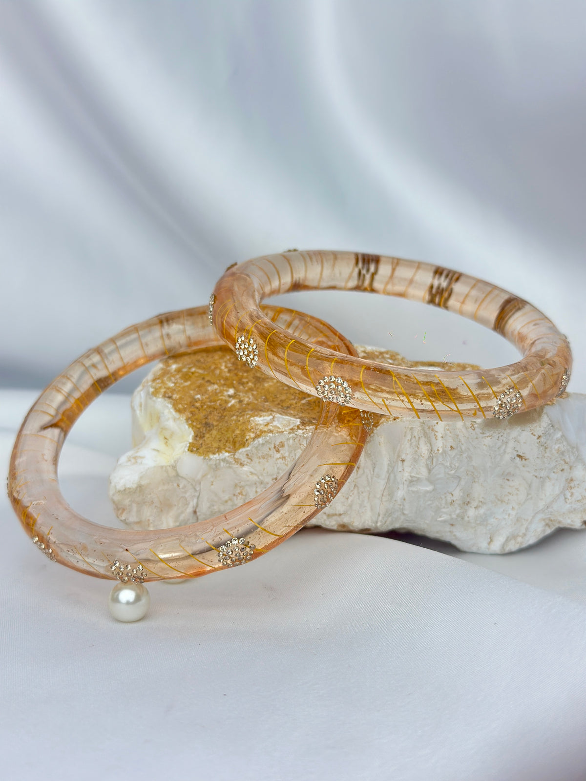 Two circular bangles with a textured, translucent amber-brown pattern and small gold-toned decorative accents, displayed on a white draped fabric backdrop with a decorative stone prop.