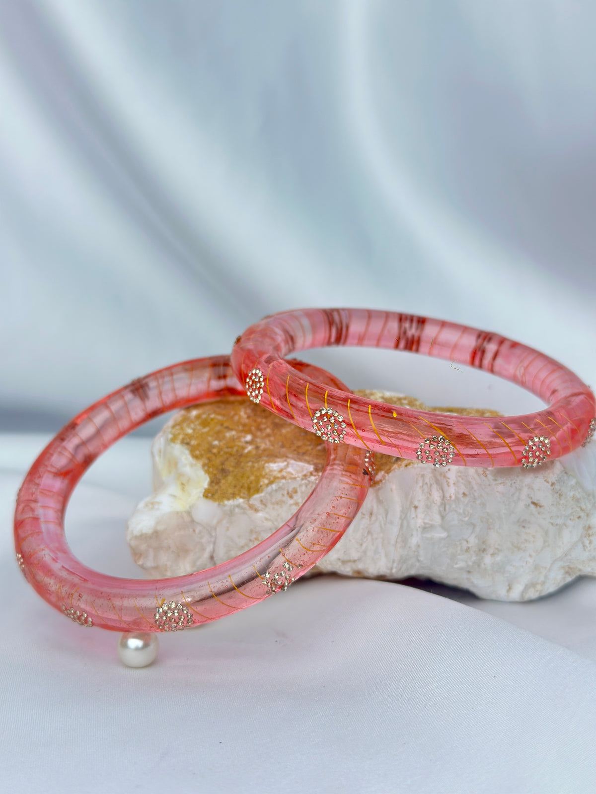 Two pink bangles with gold foil and small gold bead accents, displayed on a white stone prop against a soft white fabric background.