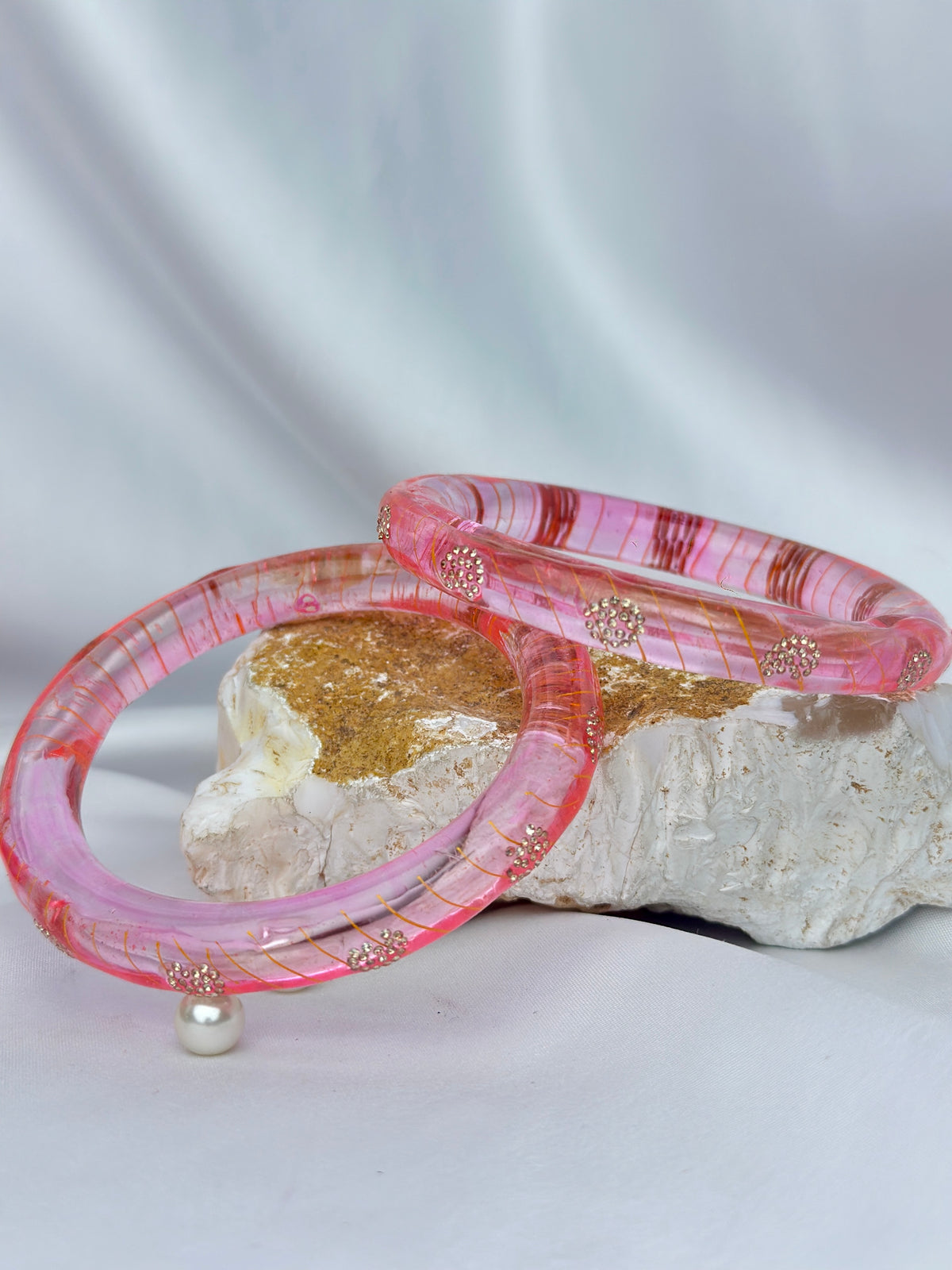Two pink marbled resin bangles with gold glitter and small gold floral accents, displayed on a white and gold decorative stone against a draped white fabric background.