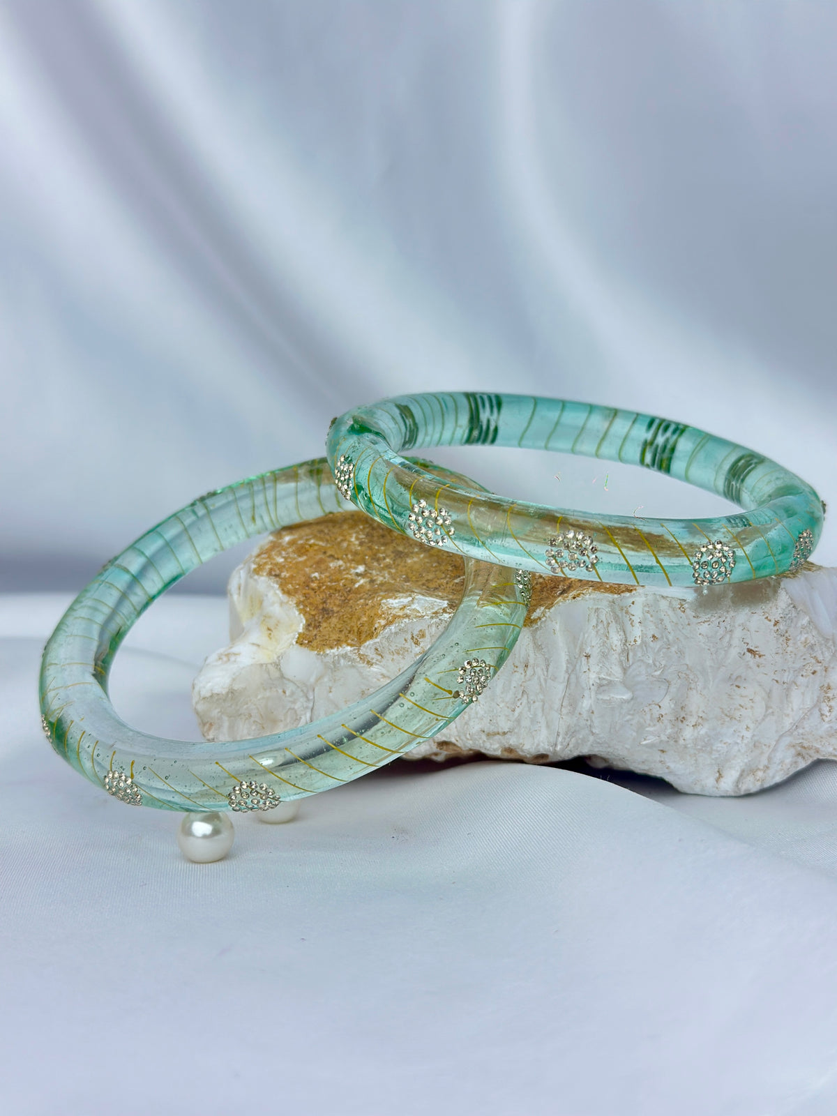 Two translucent mint green bangles with gold-toned decorative details and a subtle striped pattern, displayed on a textured stone with a small pearl accent against a draped white fabric background.