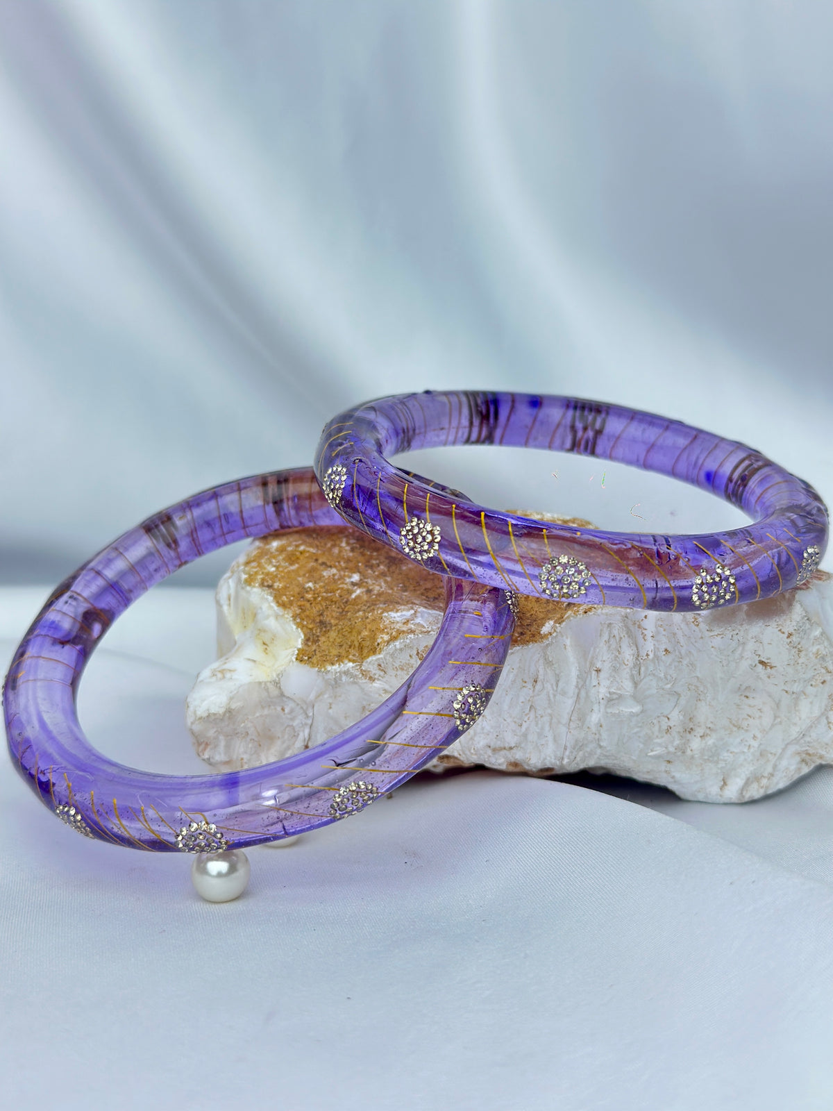 Two purple translucent bangles with gold veining and silver floral accents, displayed on a white stone prop against a soft white fabric background.