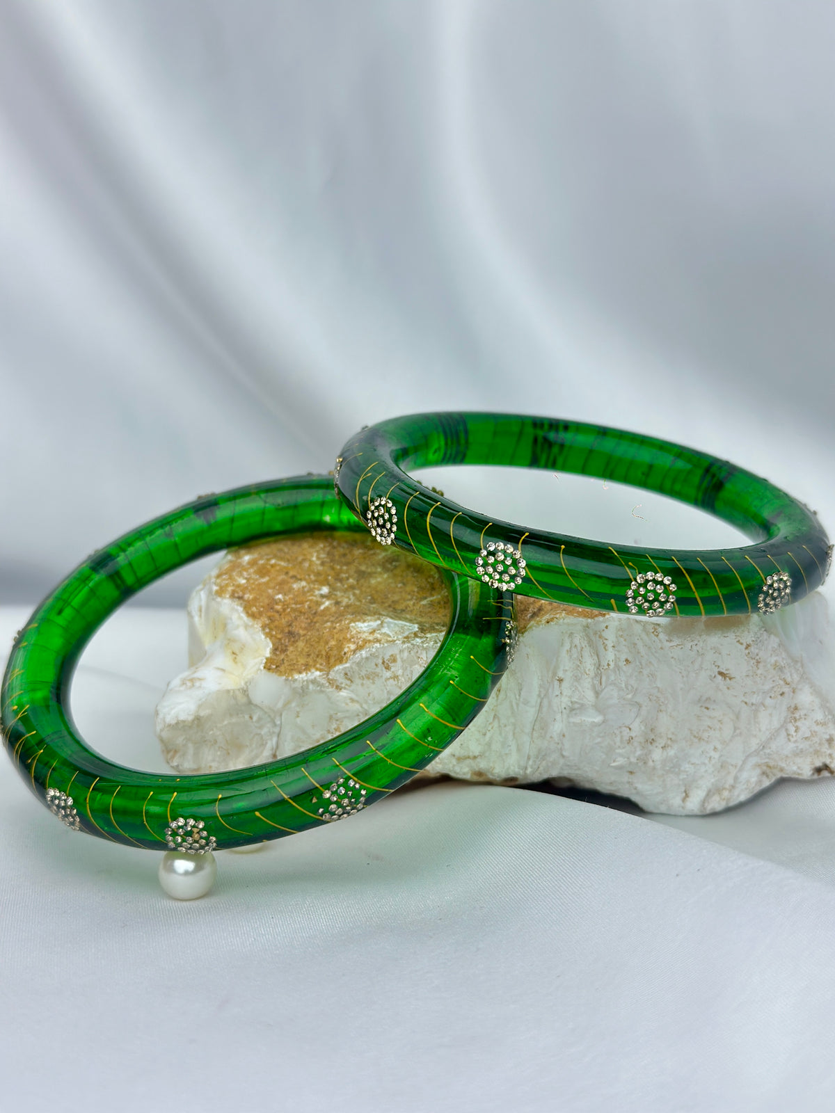 Two green bangles with gold stripes and silver floral accents, displayed on a white fabric backdrop with a decorative stone prop.