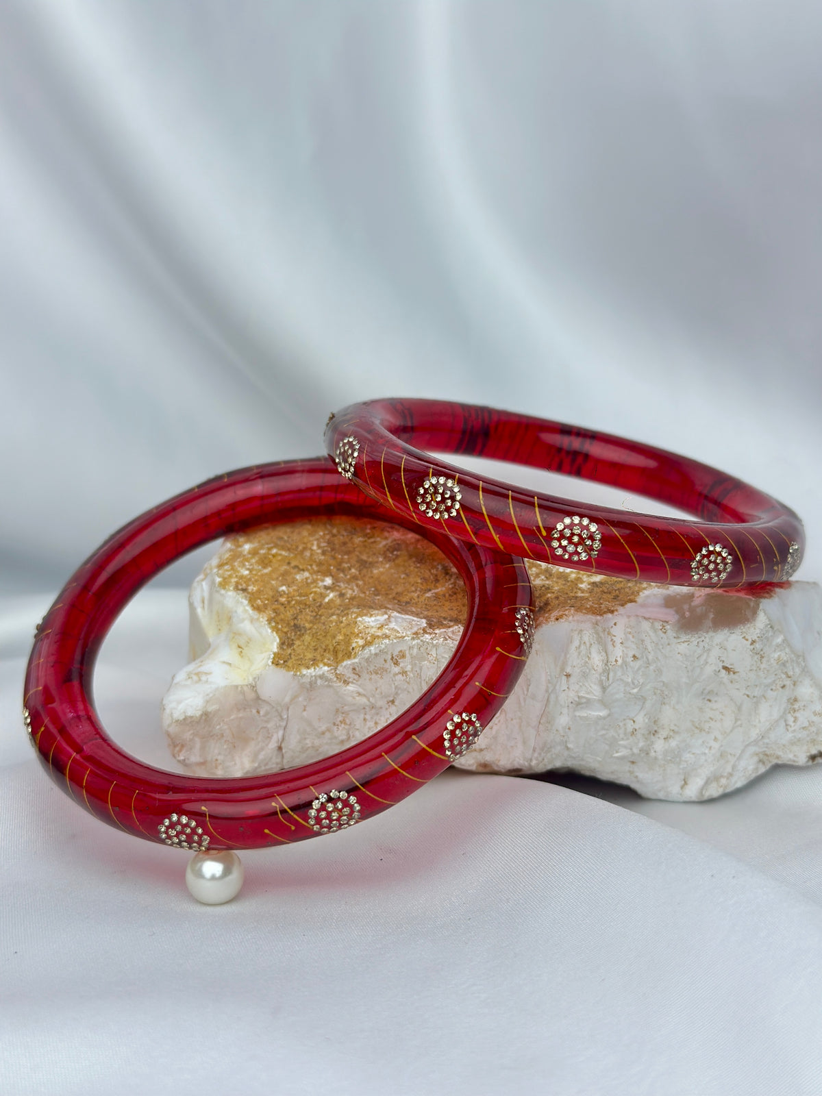 Two red bangles with gold stripes and small white stone accents, displayed on a white draped fabric backdrop with a decorative stone prop.