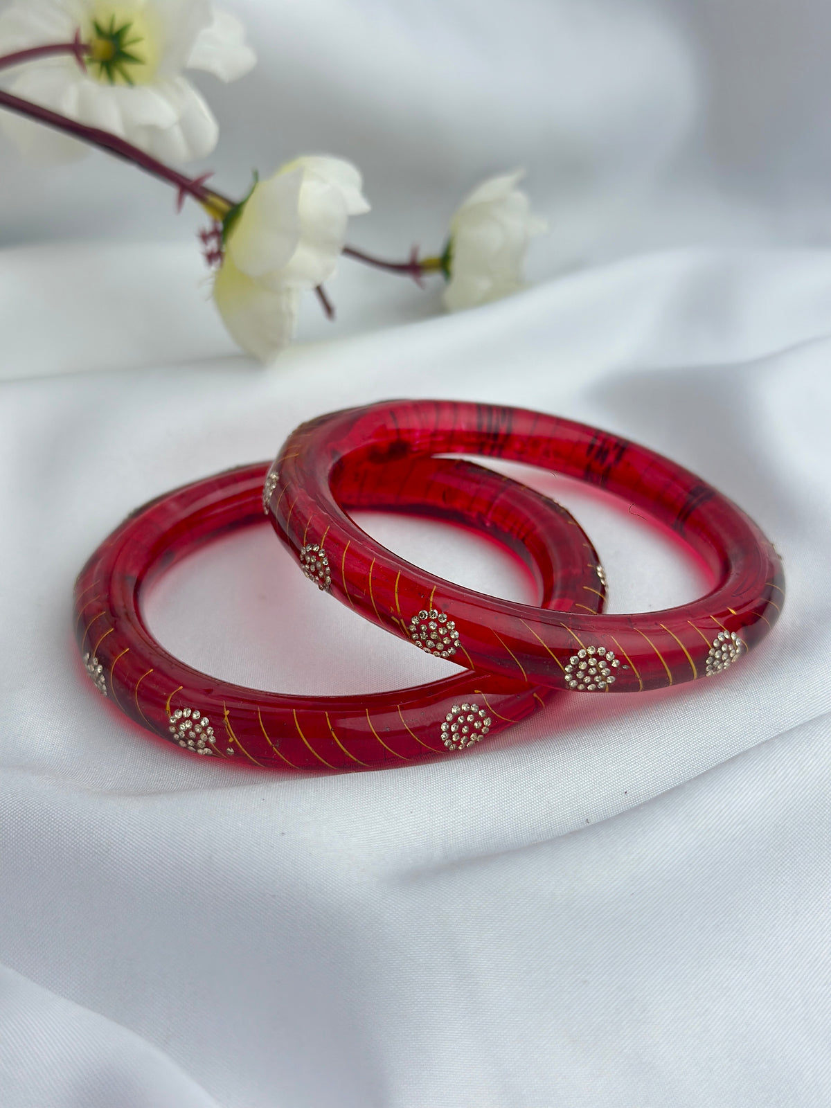 Two red glass bangles with gold stripes and small gold bead clusters, resting on a white draped fabric background with white flowers in the soft-focus background.