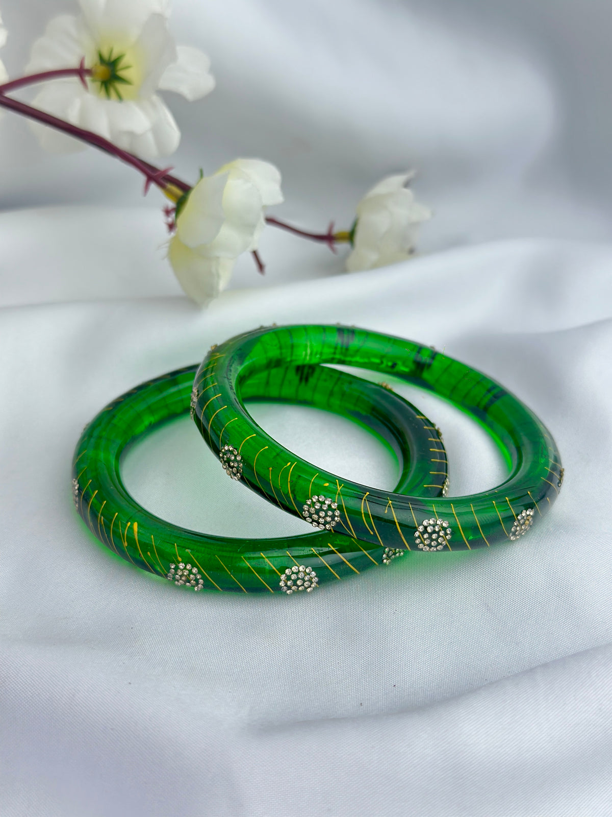 Two green bangles with gold line patterns and silver floral accents, displayed on a white fabric background with white flowers in the upper left.