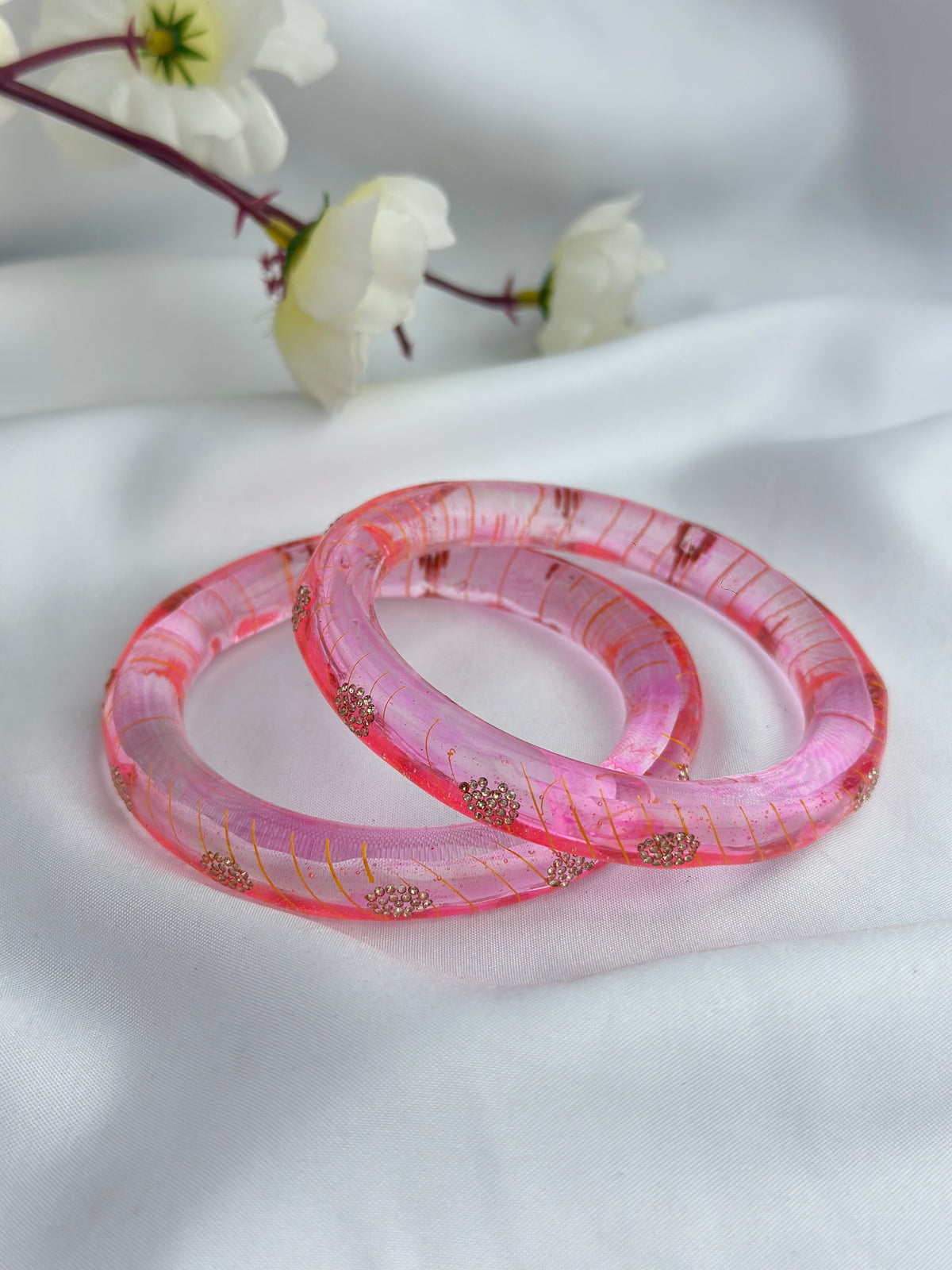 Two pink translucent bangles with gold glitter and small floral inclusions, displayed on a white fabric background with white flowers in the upper corner.