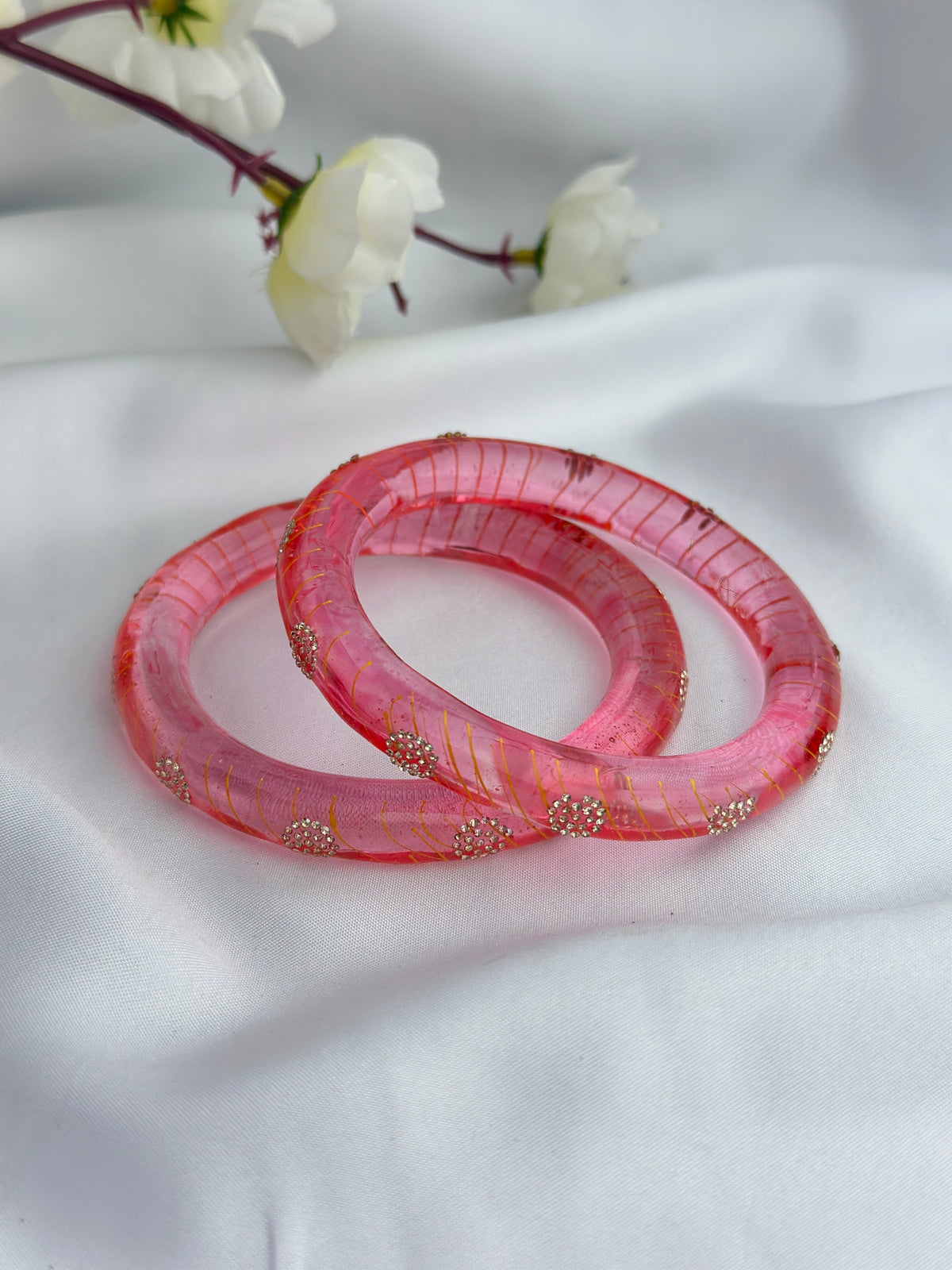 Two pink bangles with gold glitter and small metallic embellishments, resting on a white fabric background with white flowers in the upper corner.