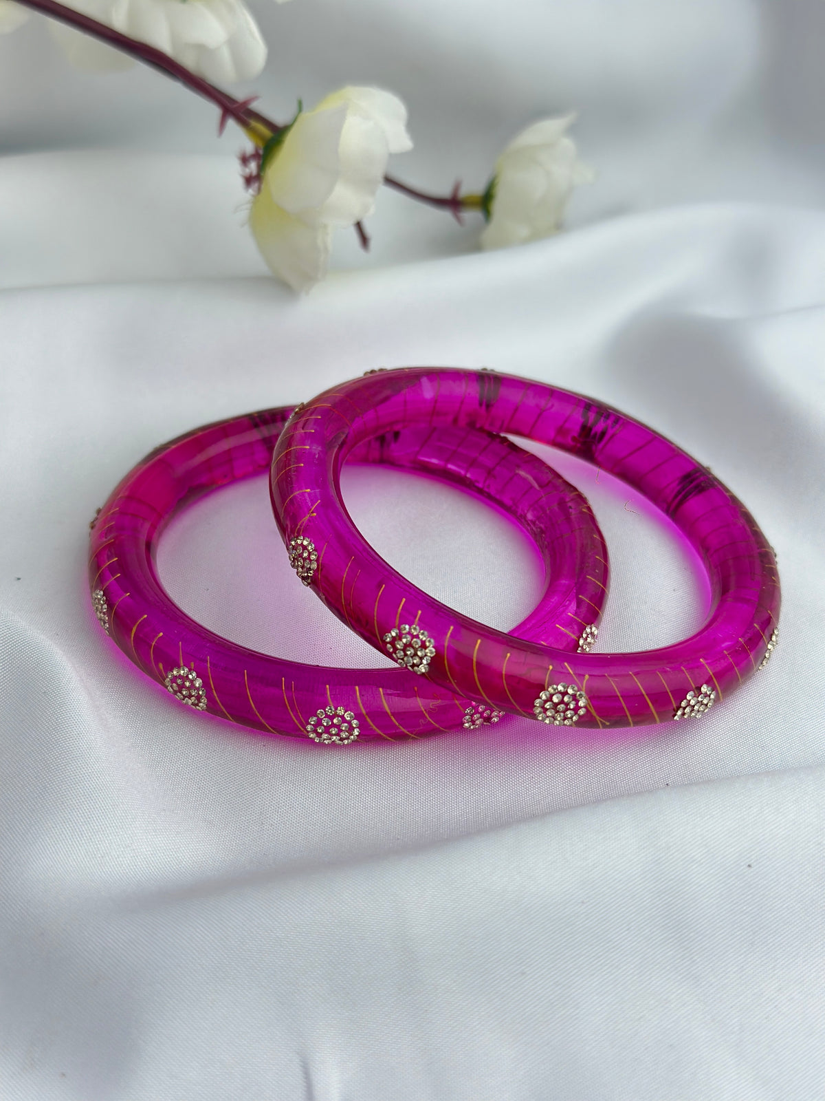Two bright magenta pink bangles with gold grid patterns and small silver rhinestone accents, displayed on a white fabric background with white flowers in the upper background.