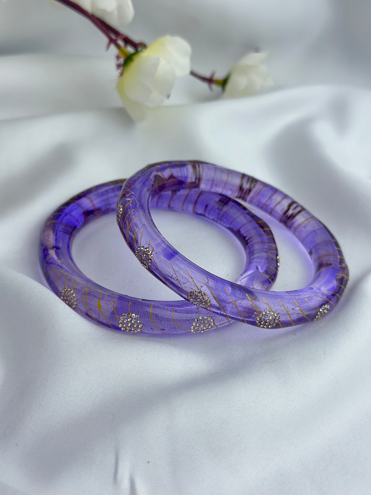 Two purple bangle bracelets with gold glitter and silver rhinestone accents, resting on a white fabric background with blurred white flowers.