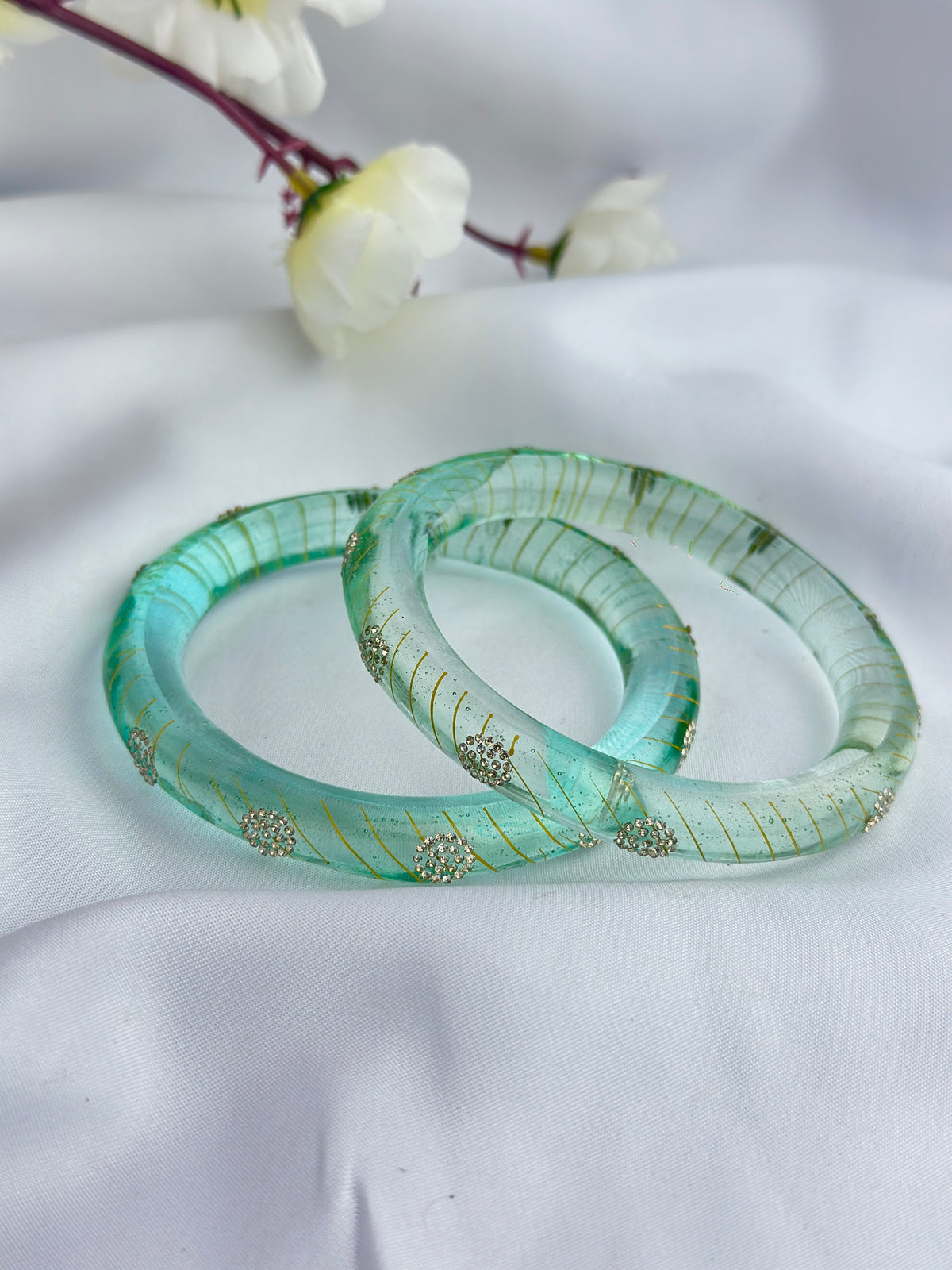 Two translucent light green bangles with gold leaf patterns and small silver bead accents, resting on a white fabric background with white flowers in the upper corner.
