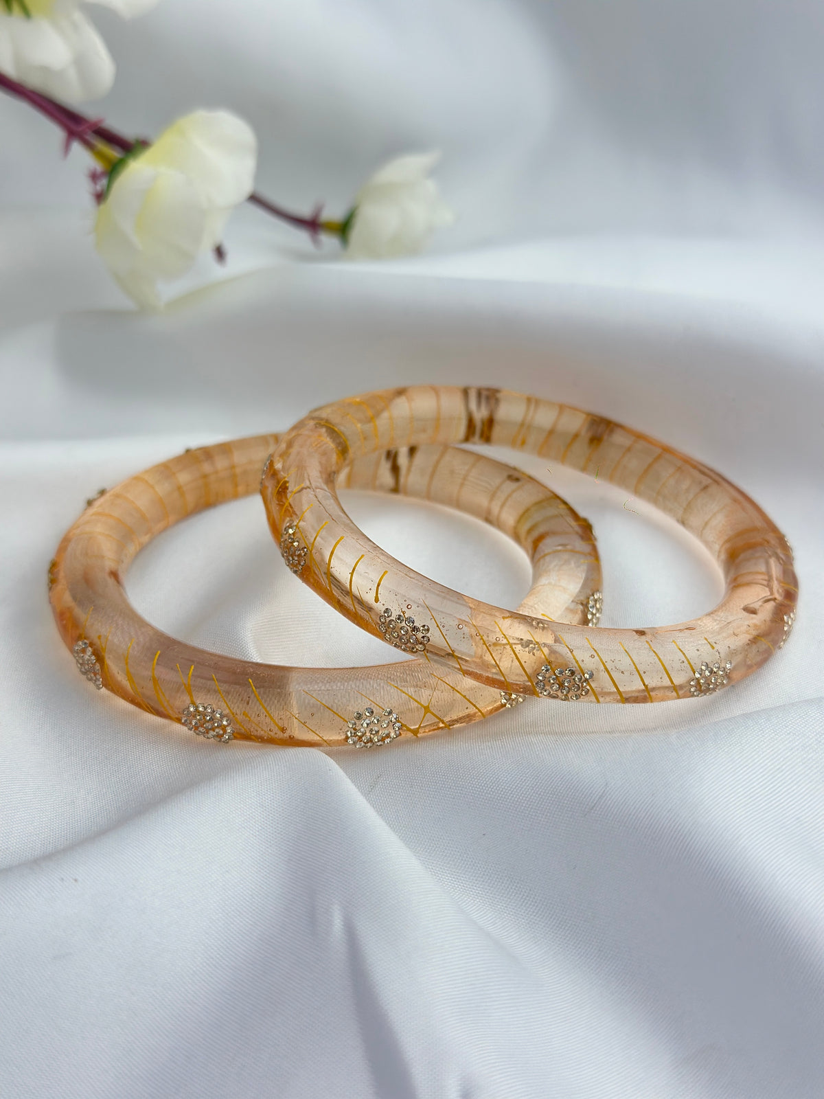 Two translucent beige bangles with gold glitter stripes and small gold floral accents, resting on a draped white fabric background with white flowers in the upper left.