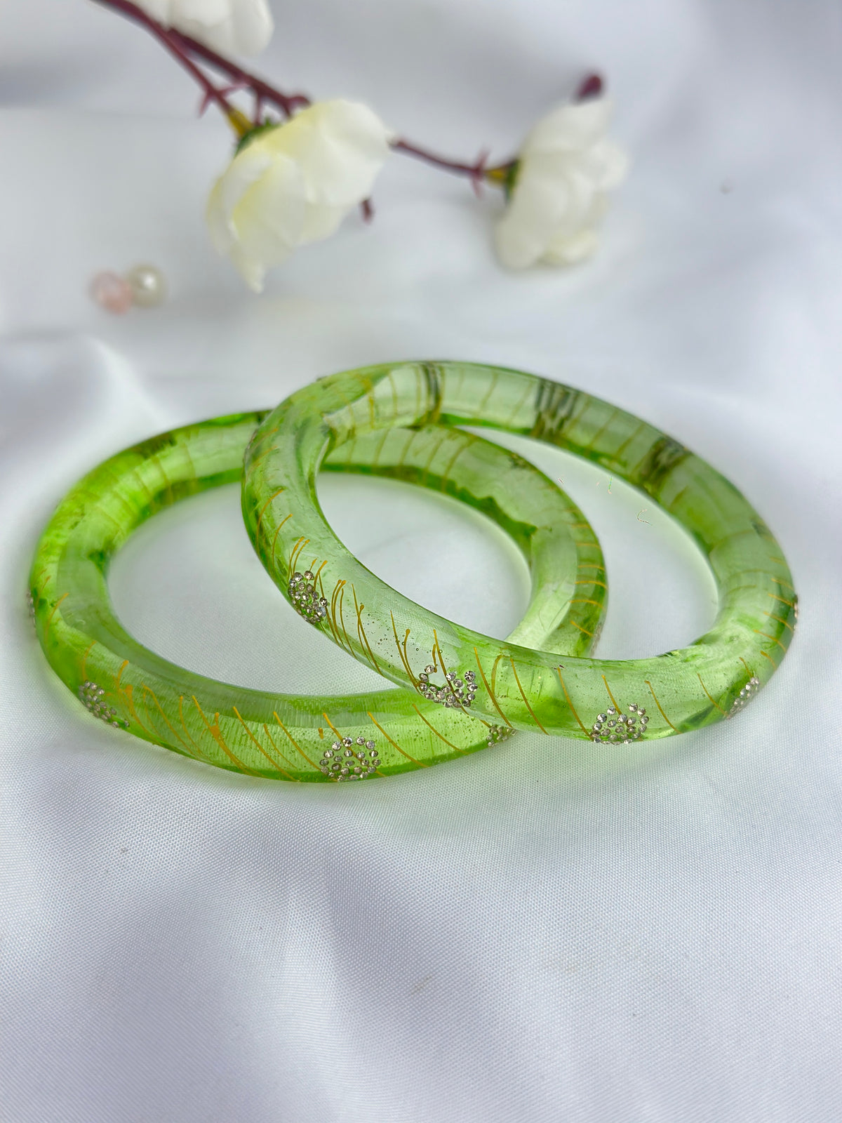 Two translucent green bangles with gold leaf inlay and small silver floral accents, displayed on a white fabric background with white flowers in the upper corner.