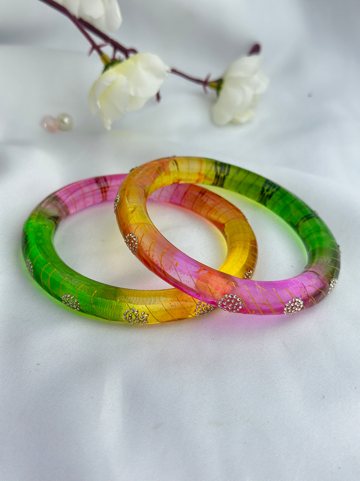 Two colorful bangle bracelets with a textured leaf inlay, featuring a green-pink gradient on one and a yellow-orange-pink gradient on the other, both adorned with small silver rhinestone accents, displayed on a white fabric background with white flowers in the background.