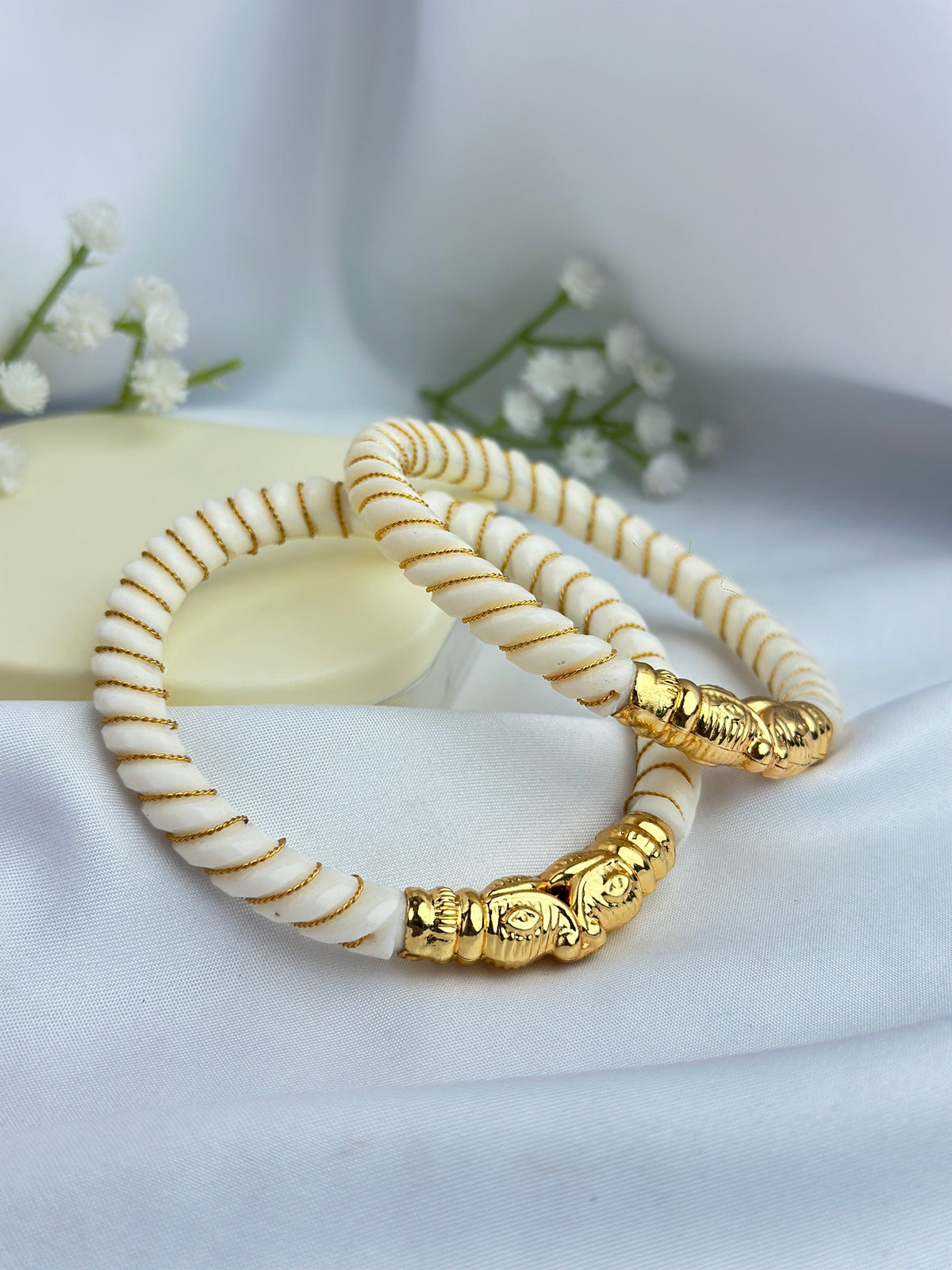 Two beaded bracelets with white beads, thin gold stripes, and gold-toned elephant-shaped charms, displayed on a soft white fabric background.