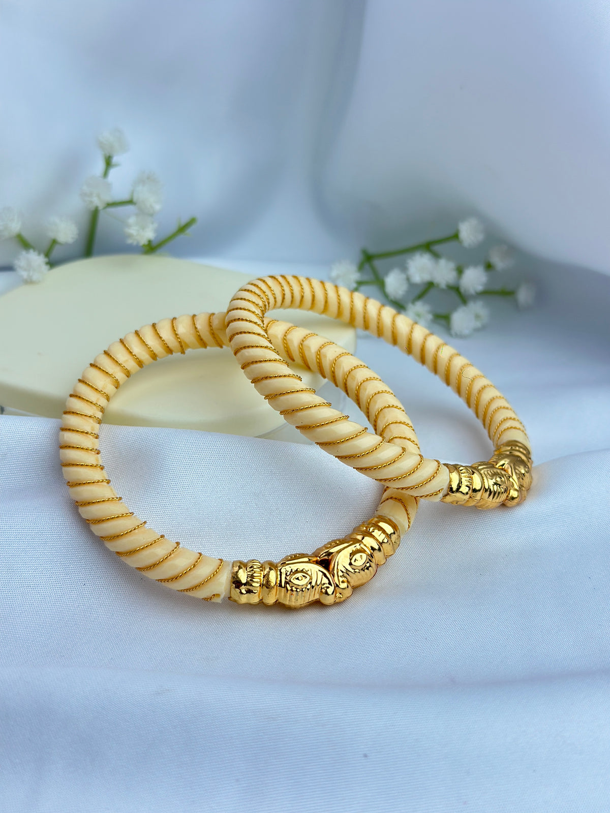 Two gold-plated beaded bracelets with textured spiral patterns and decorative elephant-shaped connectors, displayed on a soft white fabric background with small white flowers.