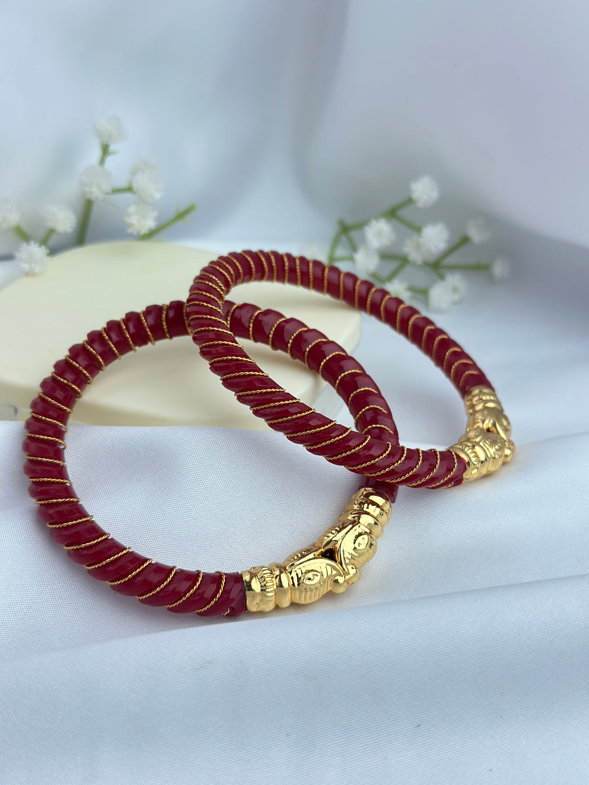 Two red braided bracelet-style accessories with gold-toned decorative ends, arranged on a soft white fabric background with small white flowers and a cream block behind them.