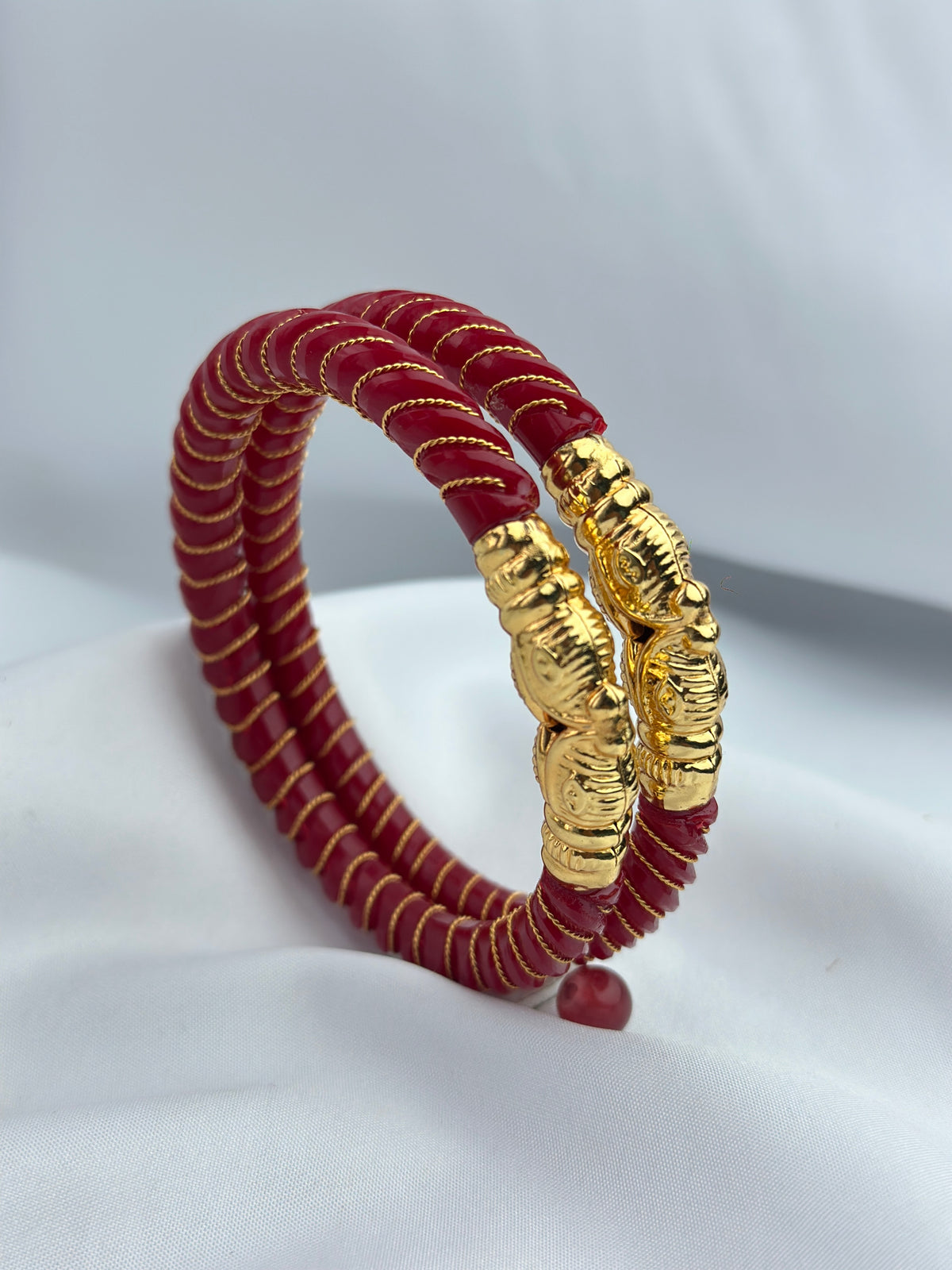 A red beaded bracelet with gold accents, featuring two textured gold elephant-shaped clasps and gold thread wrapping, displayed on a white fabric background.