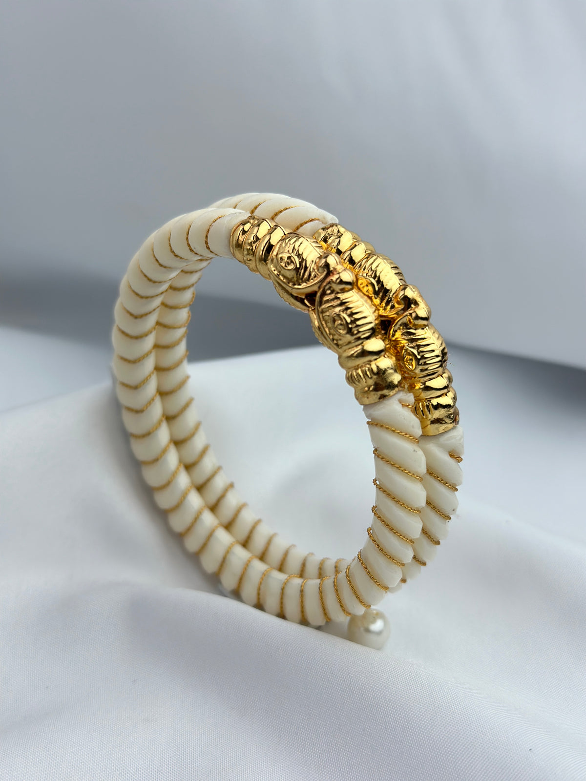 A beaded hinged bracelet featuring cream-colored round beads with gold-toned ridges and a textured gold-toned decorative clasp, displayed on a soft white fabric background.
